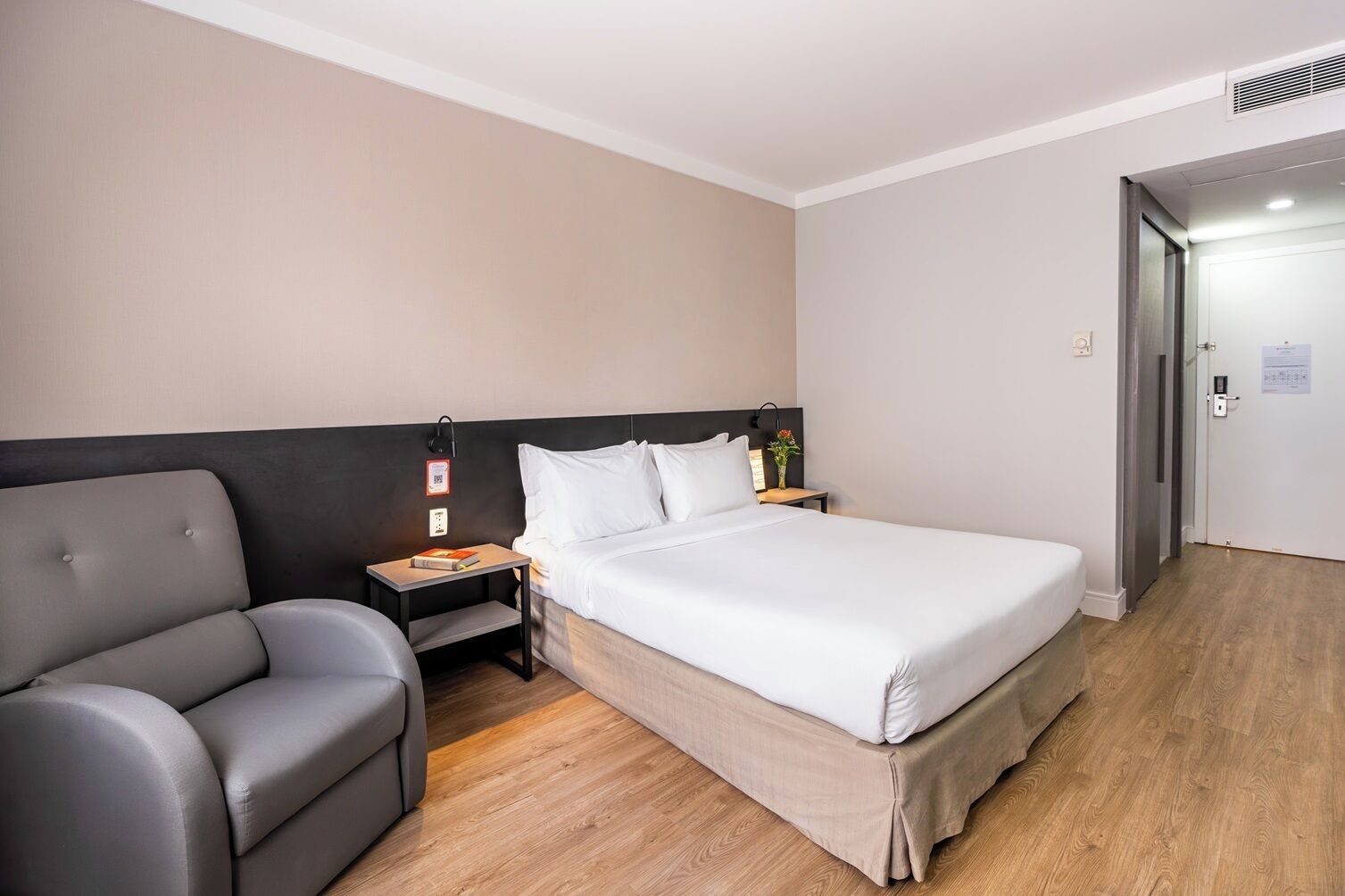 A hotel room with a bed, a gray armchair, a bedside table with a lamp, and a door with a lock.