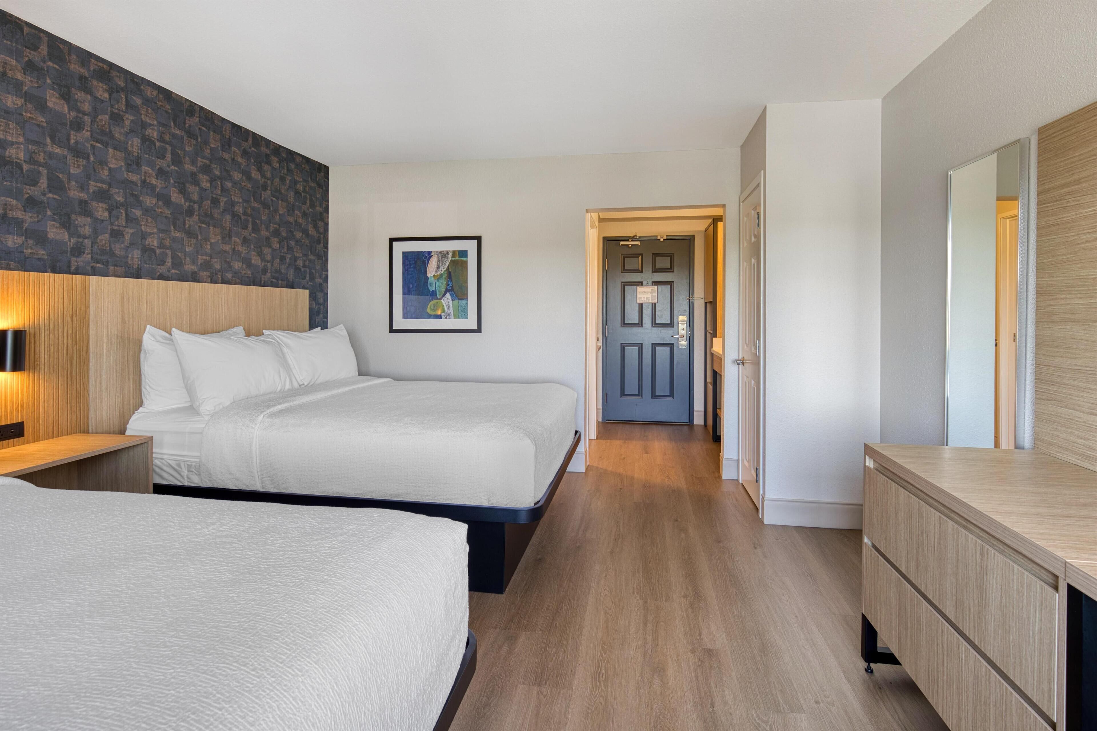 Habitación de hotel con dos camas, un cabecero de madera, una mesita de noche con lámpara, un aparador y un cuadro enmarcado en la pared.