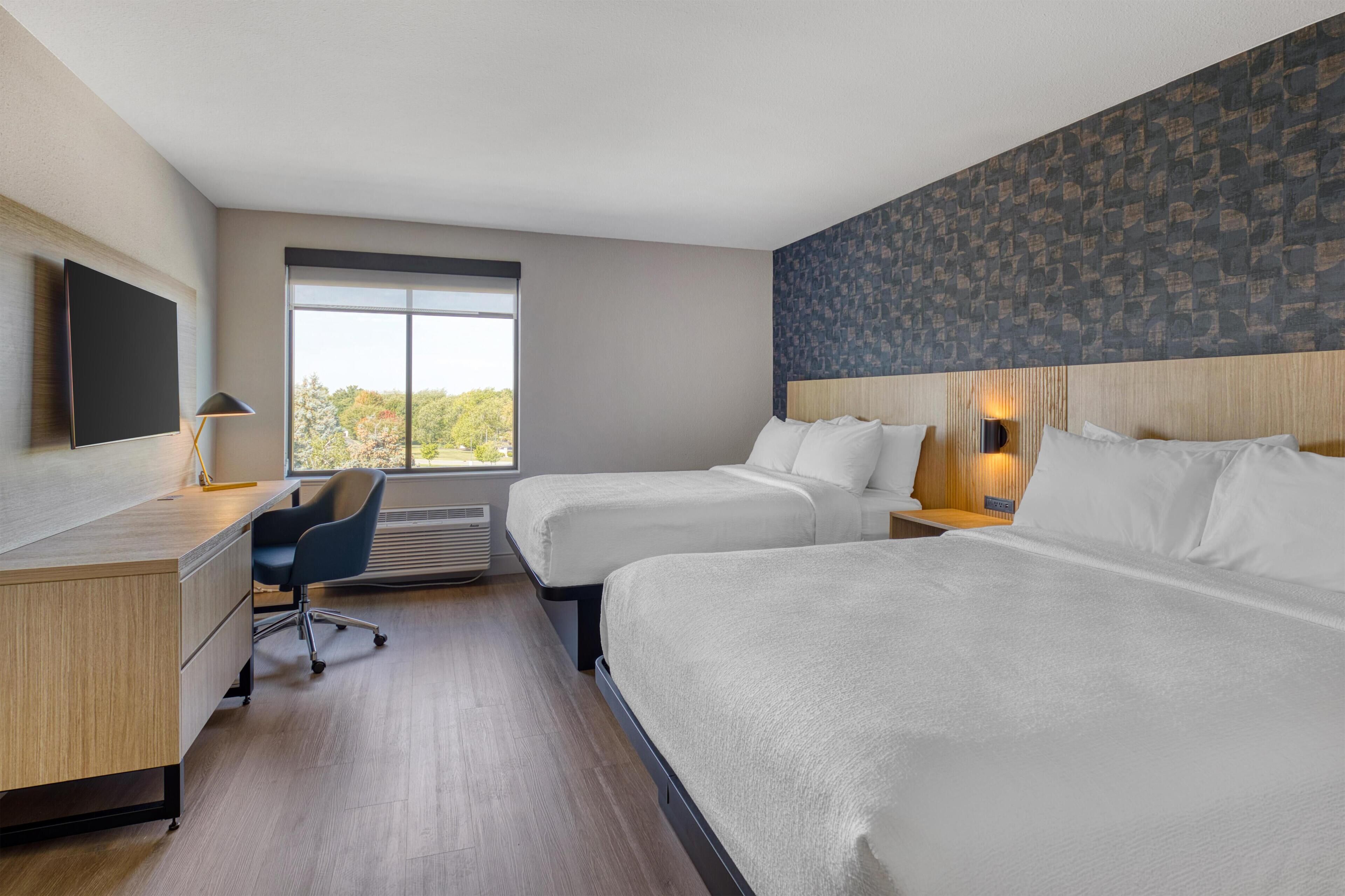 Una habitación de hotel con dos camas, un escritorio, una silla, un televisor y un ventanal grande.