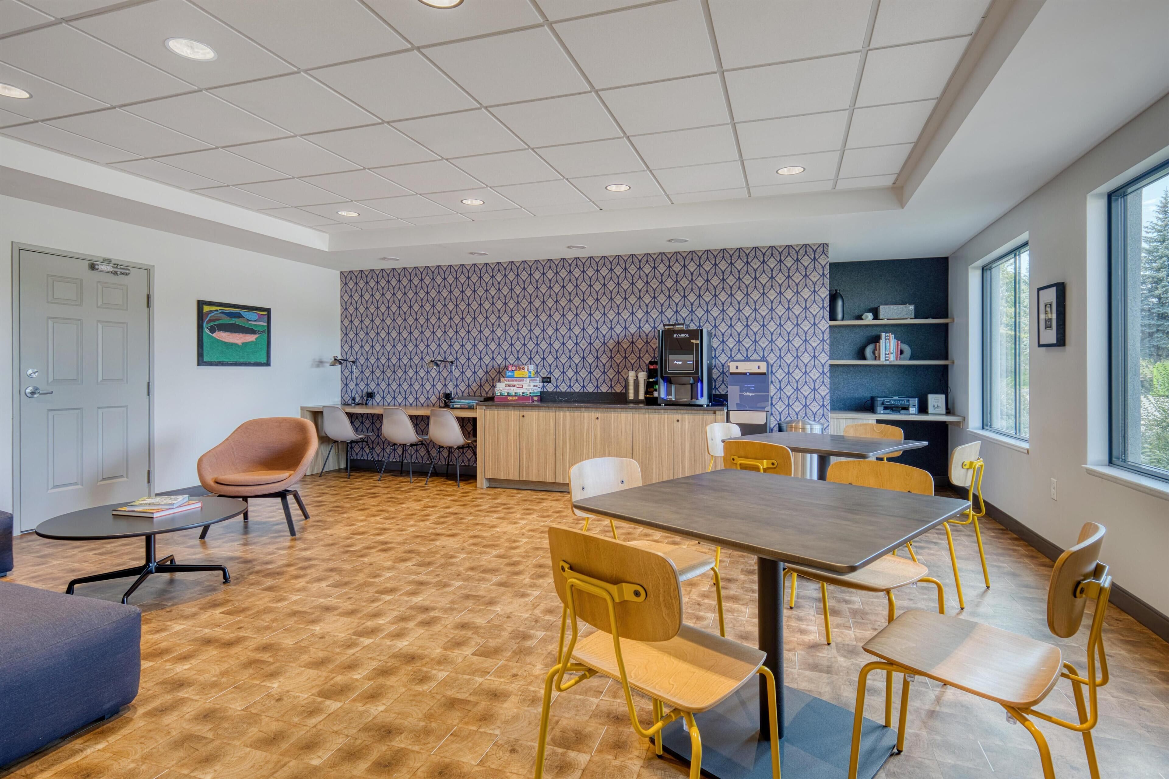 A modern meeting room with a wooden floor, a large table, and yellow chairs. There is a reception desk with a patterned wallpaper and a computer.