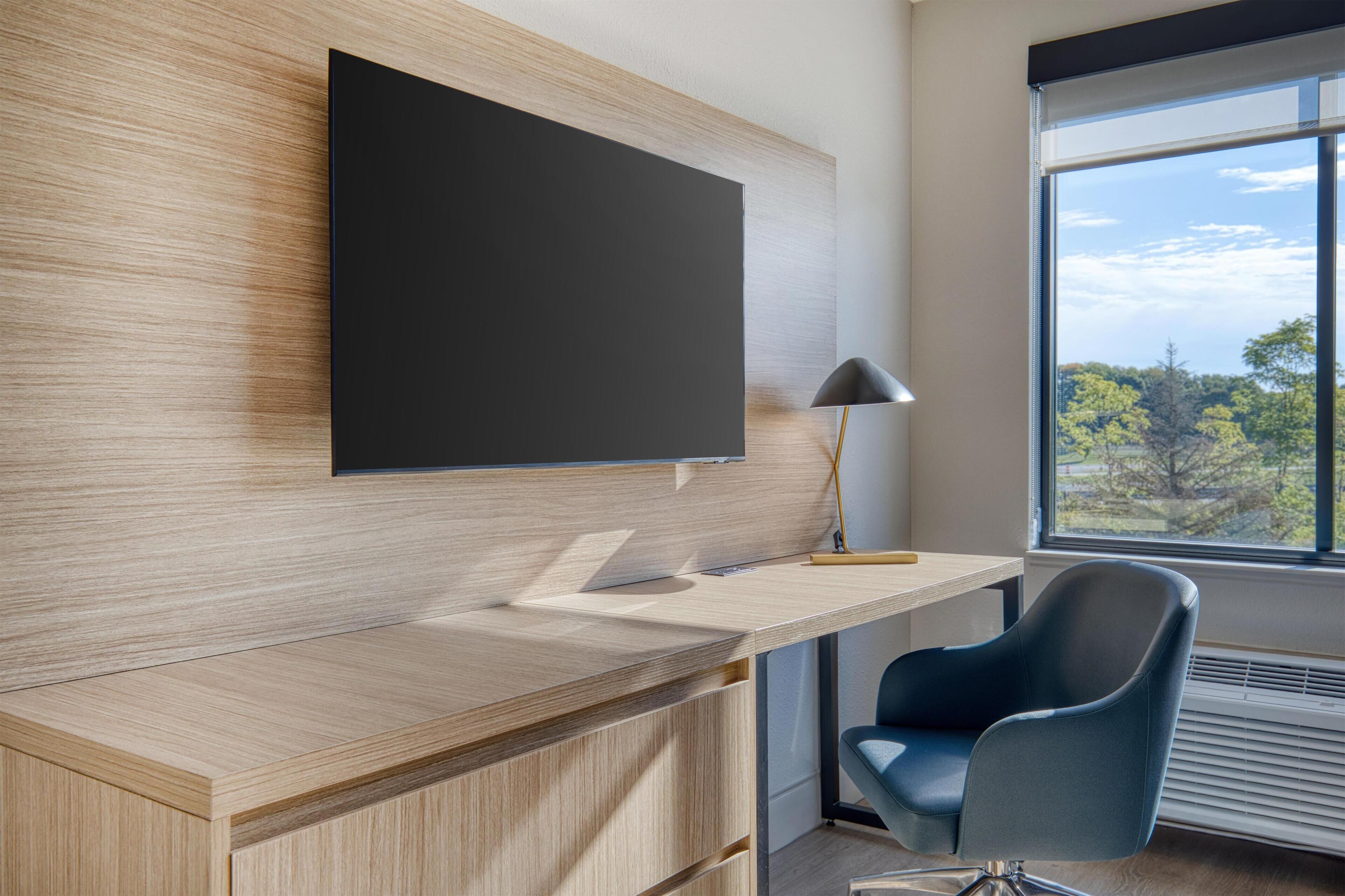 A modern office space with a wooden desk, a blue chair, a mounted flat-screen TV, and a window with a view of trees.