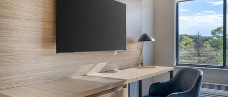 A modern office space with a wooden desk, a blue chair, a mounted flat-screen TV, and a window with a view of trees.