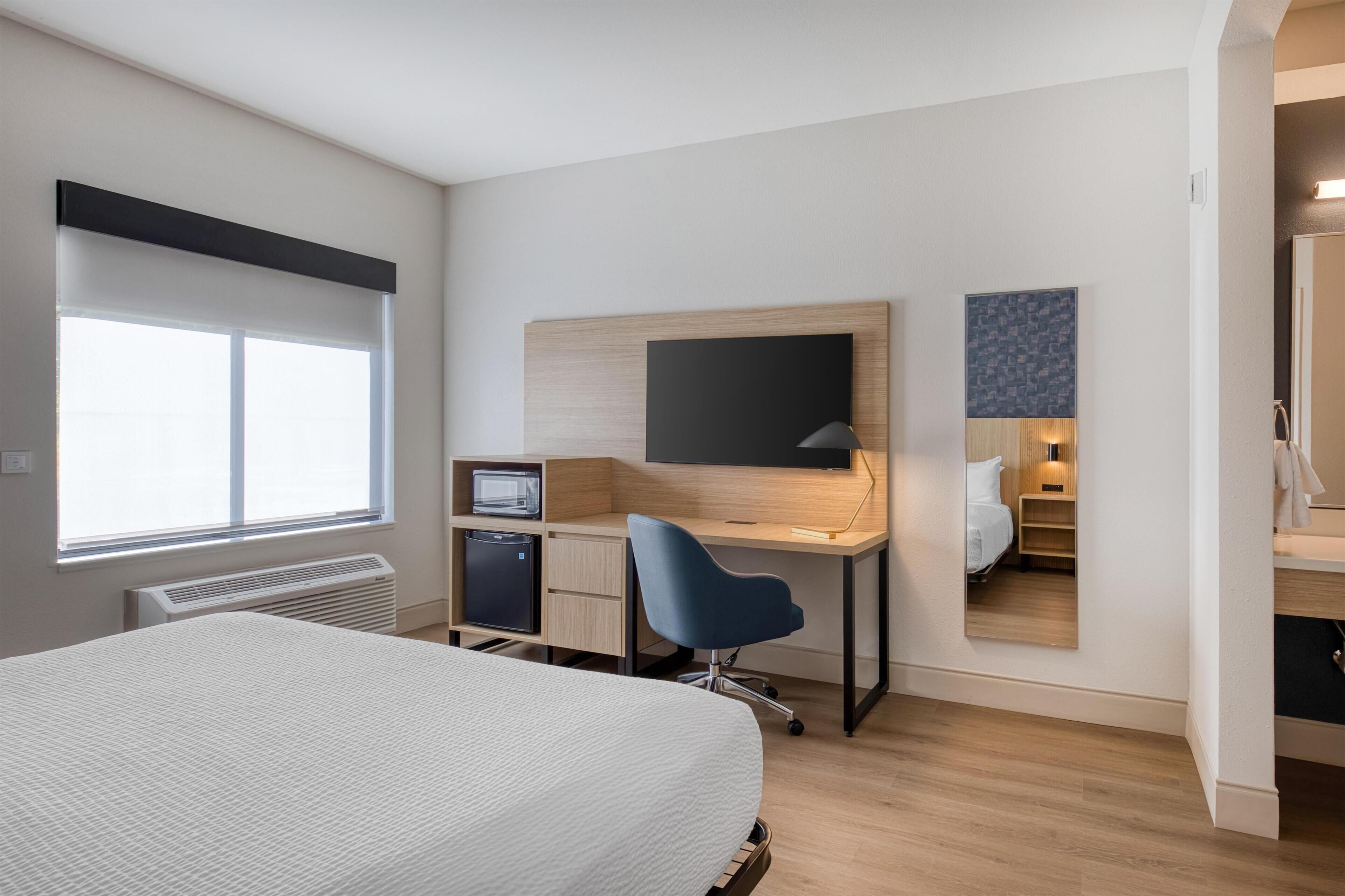 Una habitación de hotel moderna con una cama grande, un escritorio con computadora, un televisor de pantalla plana y una pequeña kitchenette.