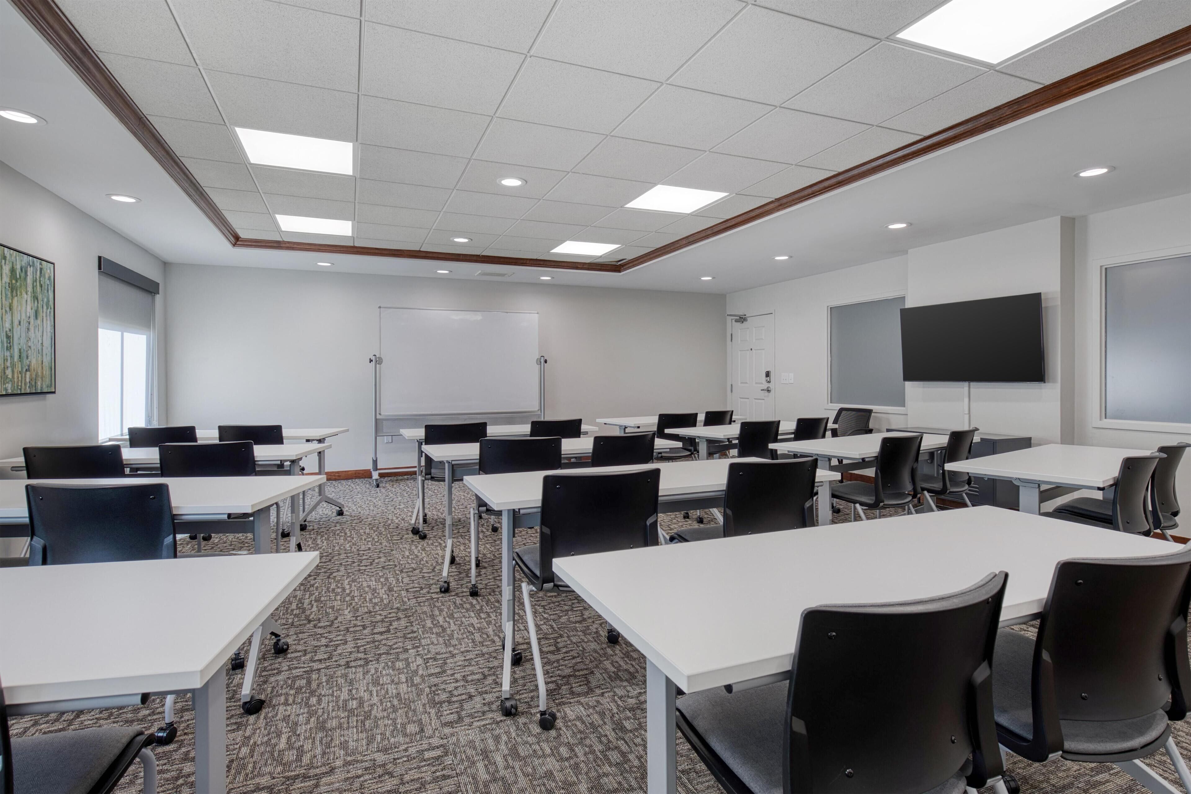 A conference room with white tables, black chairs, a whiteboard, a flat-screen TV, and a carpeted floor.