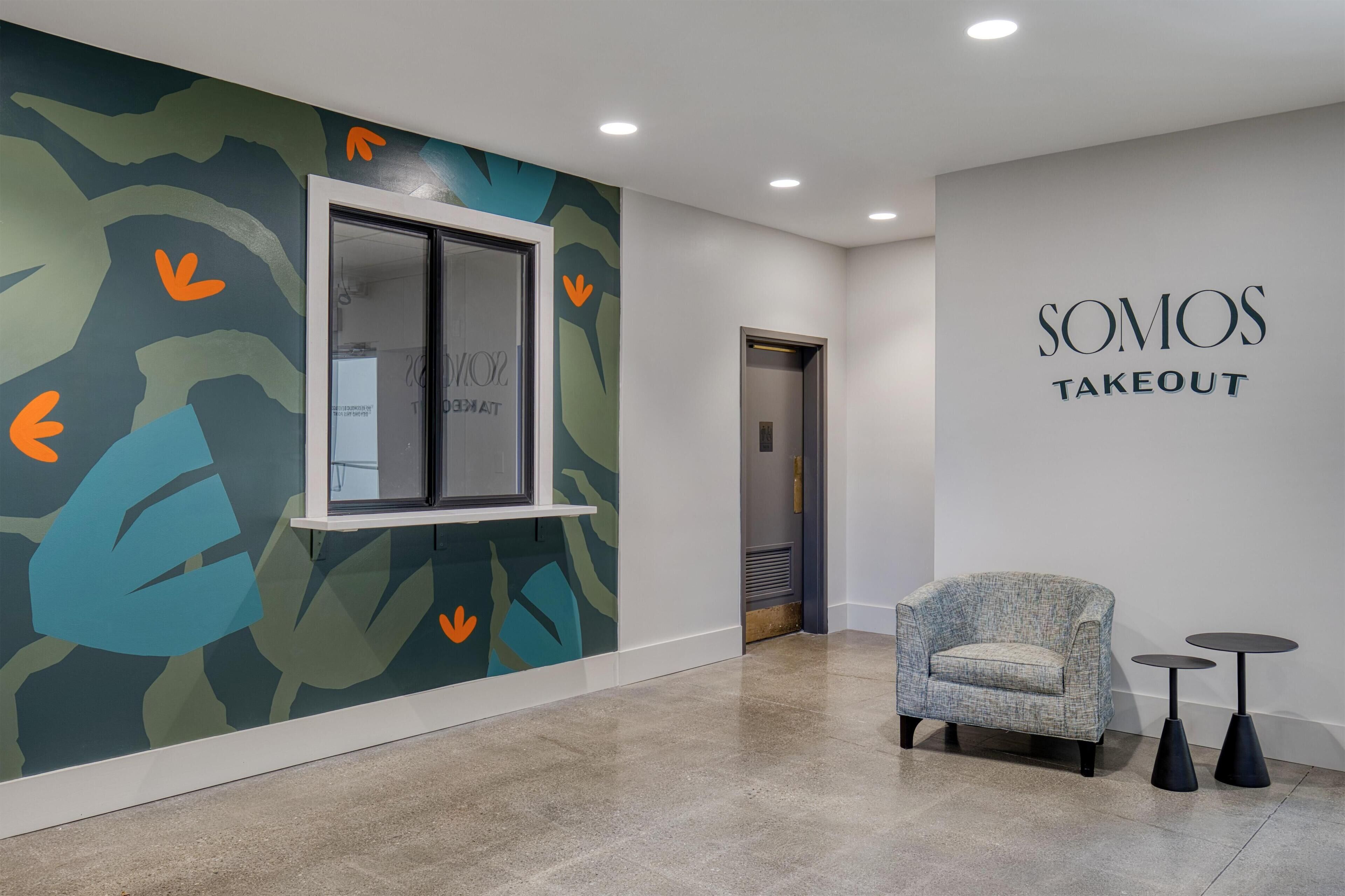 A modern interior with a wall-mounted window, a seating area with a chair and two small tables, and a wall with the text 'SOMOS TAKEOUT'.