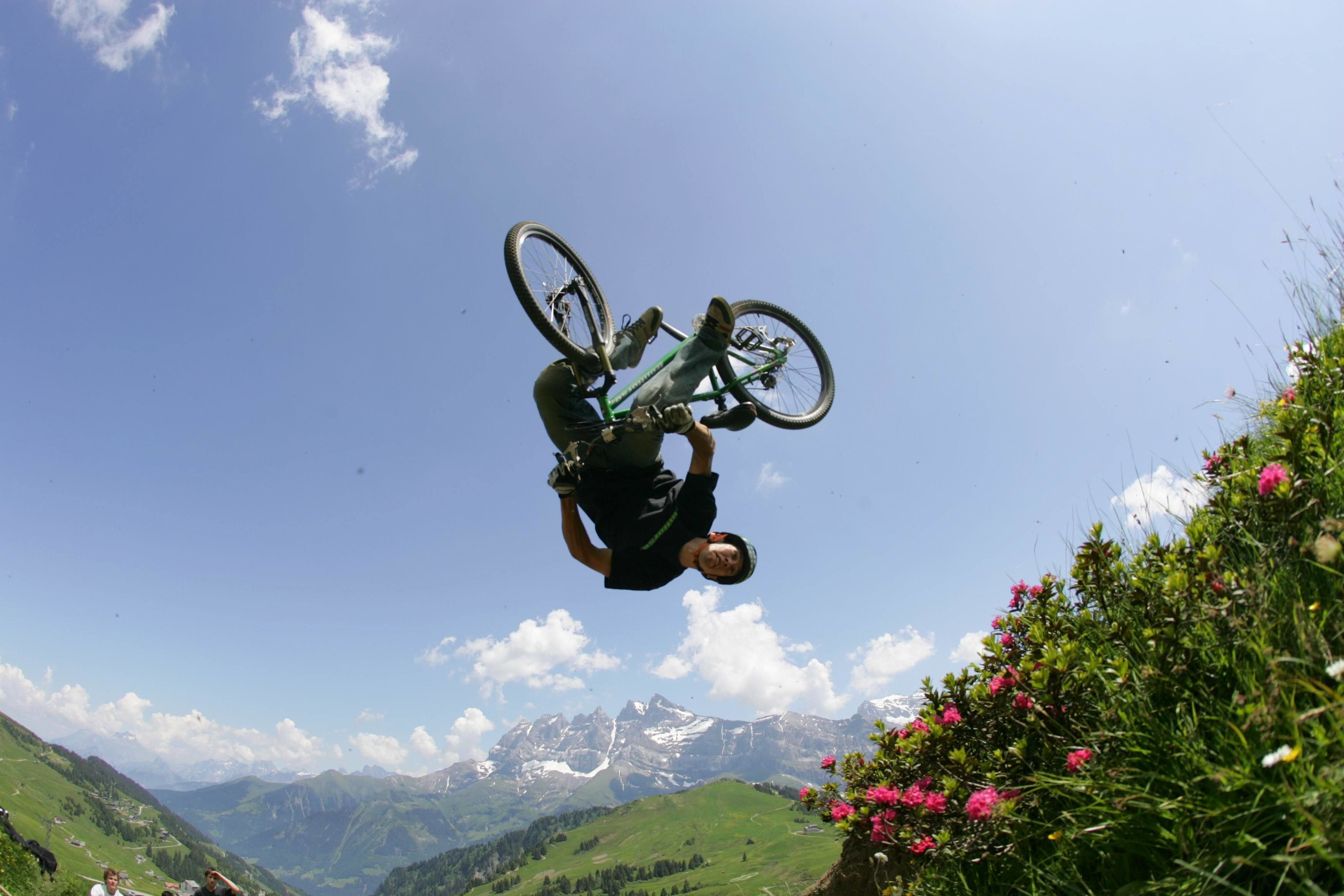 Una persona realiza una acrobacia en bicicleta en una zona montañosa con cumbres visibles y vegetación.