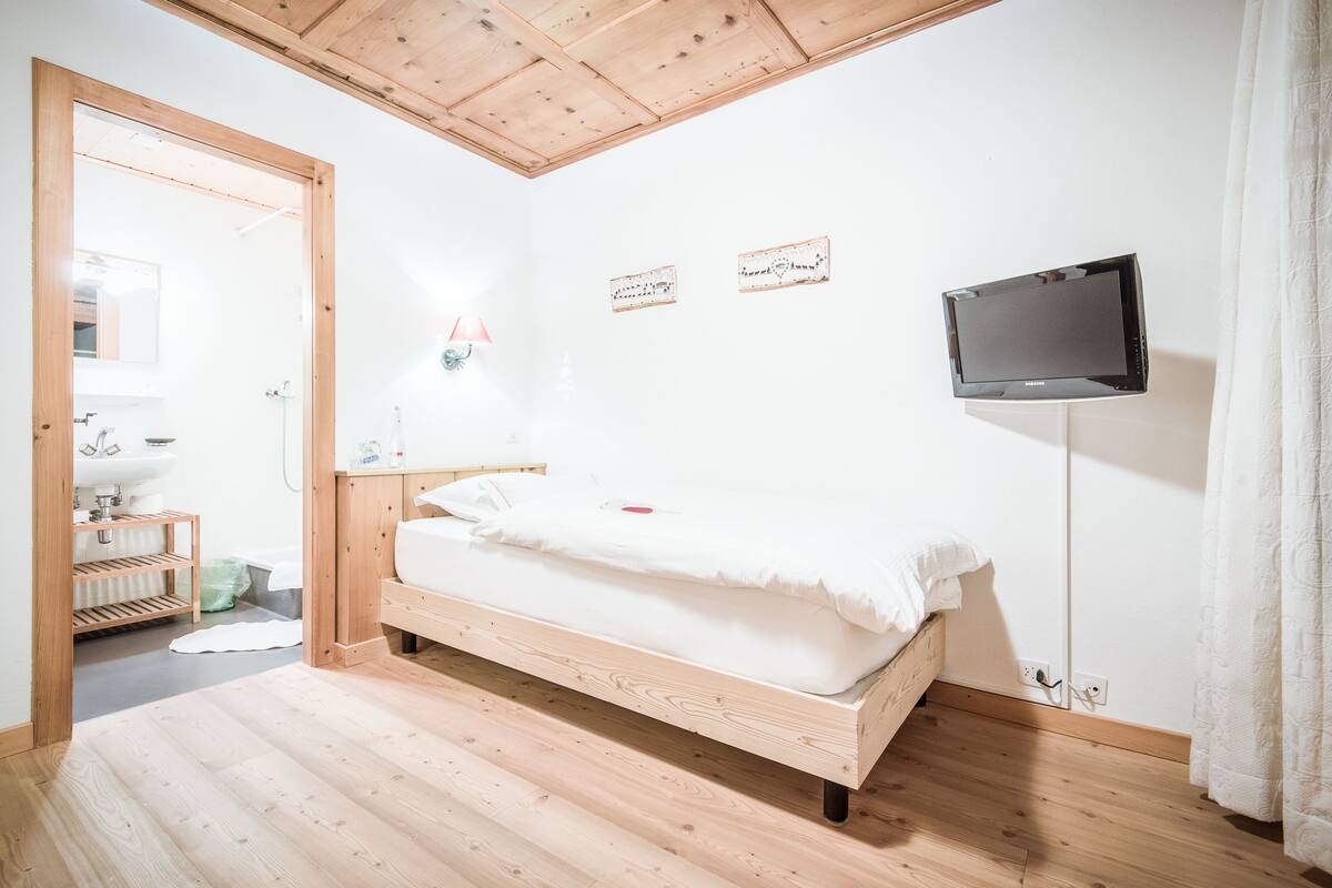 Ein Schlafzimmer mit einem hölzernen Bett, einem an der Wand montierten Fernseher und einem durch eine offene Tür einsehbaren Badezimmer.