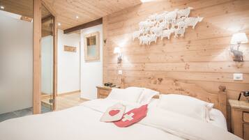 Un dormitorio con techo de madera, cama con ropa de cama blanca, mesita de noche con lámpara y pared decorada con recortes de ovejas.