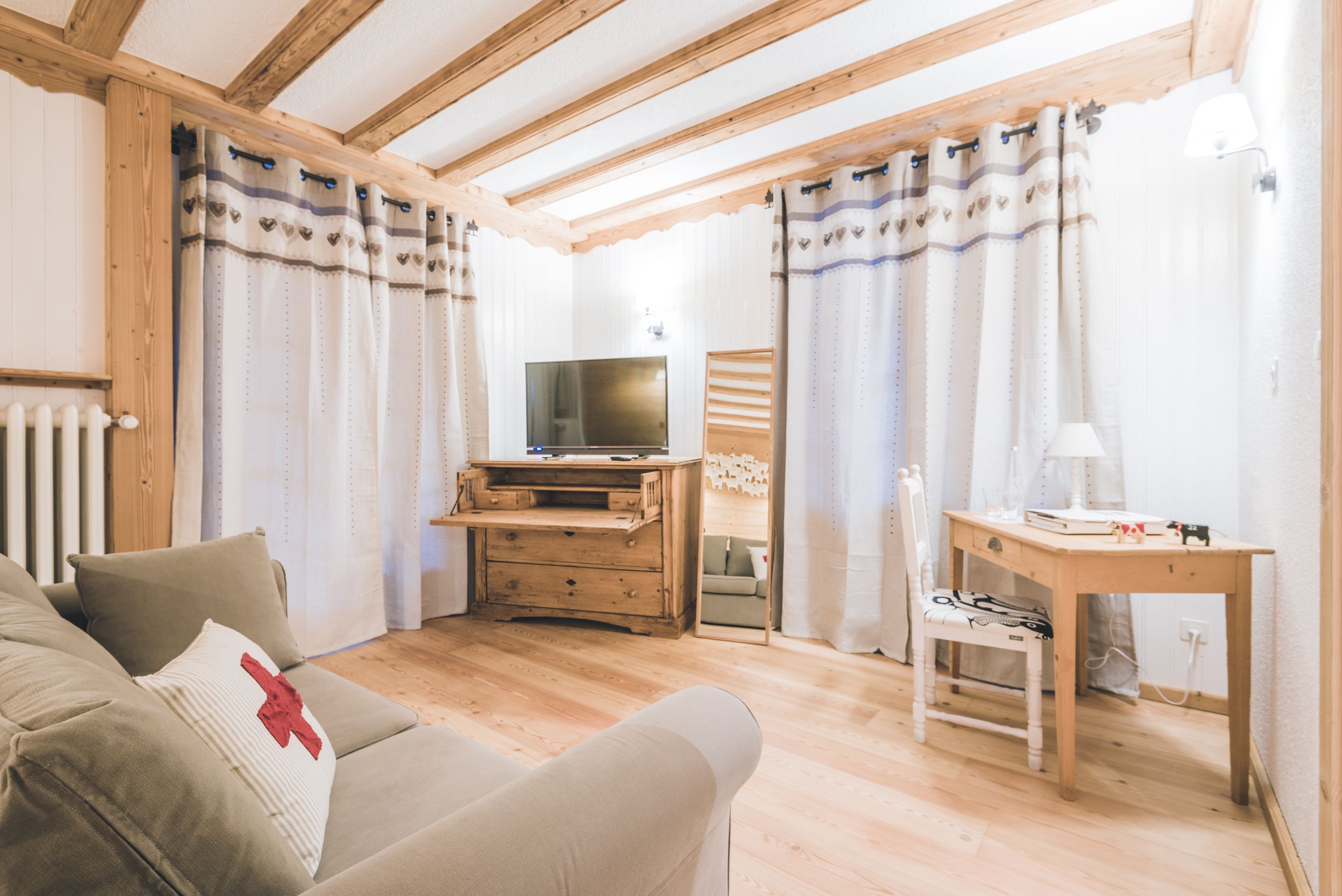 A cozy living room with a wooden ceiling, a television on a wooden stand, a sofa with a red pillow, and a small table with a lamp.