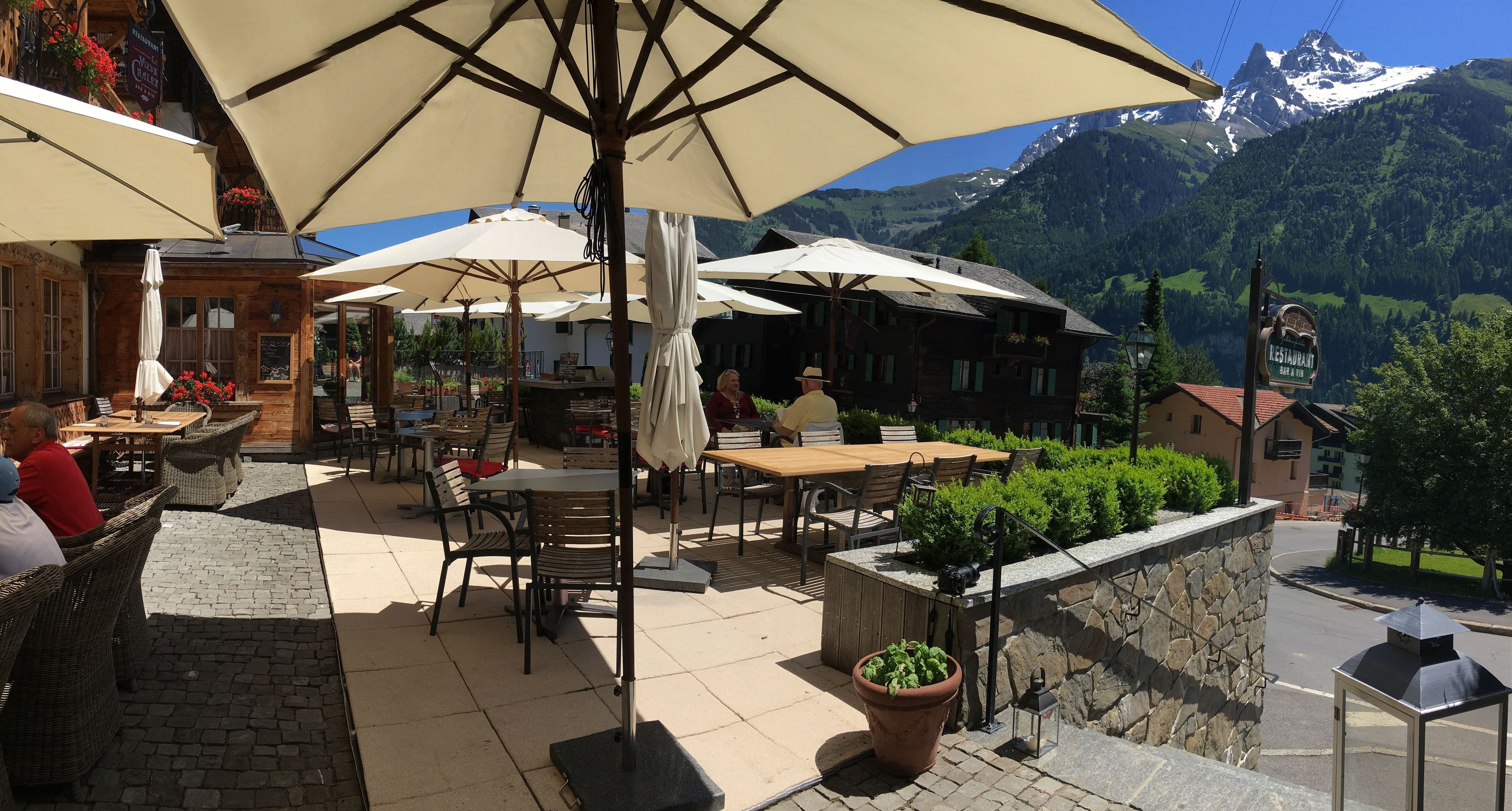 Un café en plein air, avec des tables et des chaises disposées sous de grands parasols, le tout sur fond de montagnes et d’un ciel dégagé.