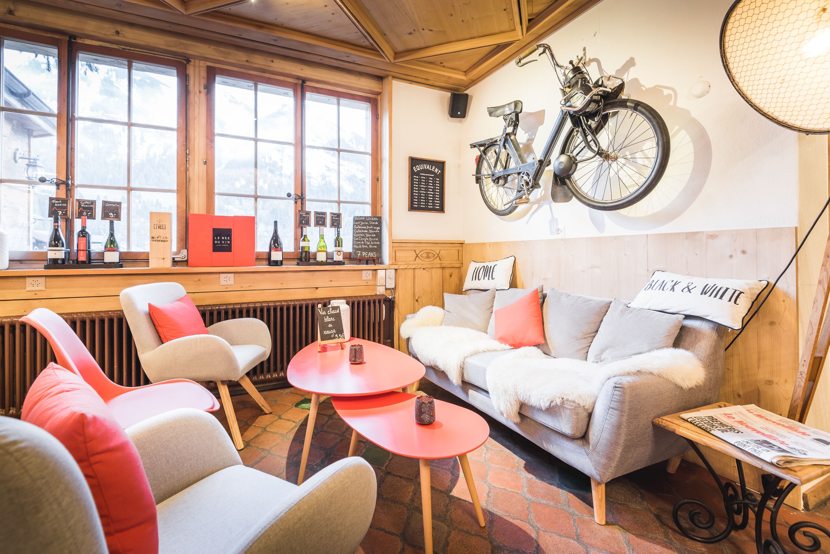 Een gezellig interieur met een houten plafond, een bank met 'BLACK & WHITE' kussens, een fiets aan de muur en een barhoek met wijnflessen.