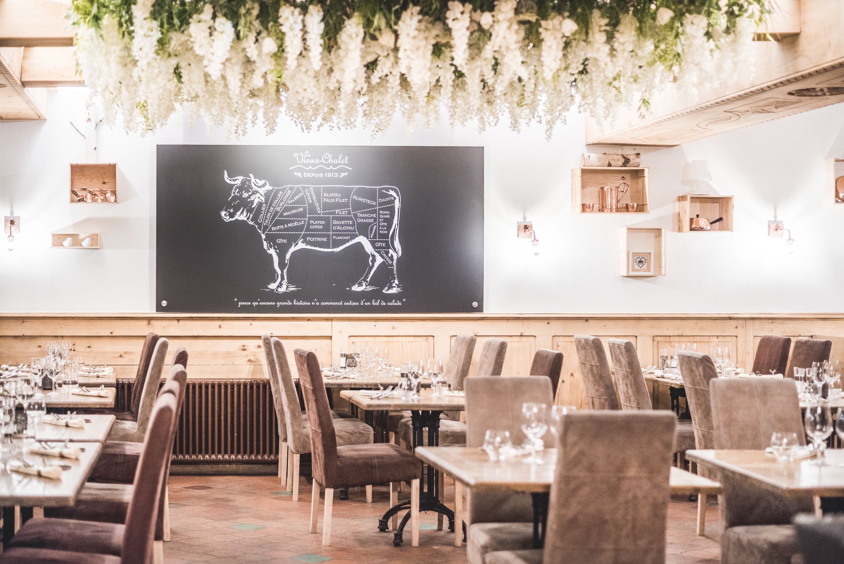 Ein Restaurant mit einer Tafel, die eine Bullenillustration und Text enthält, Holztische und -stühle sowie hängende weiße Blumen.