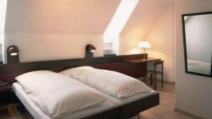 Un lit bien fait dans une pièce dont le plafond est en pente, un bureau avec une lampe et un grand miroir accroché au mur.