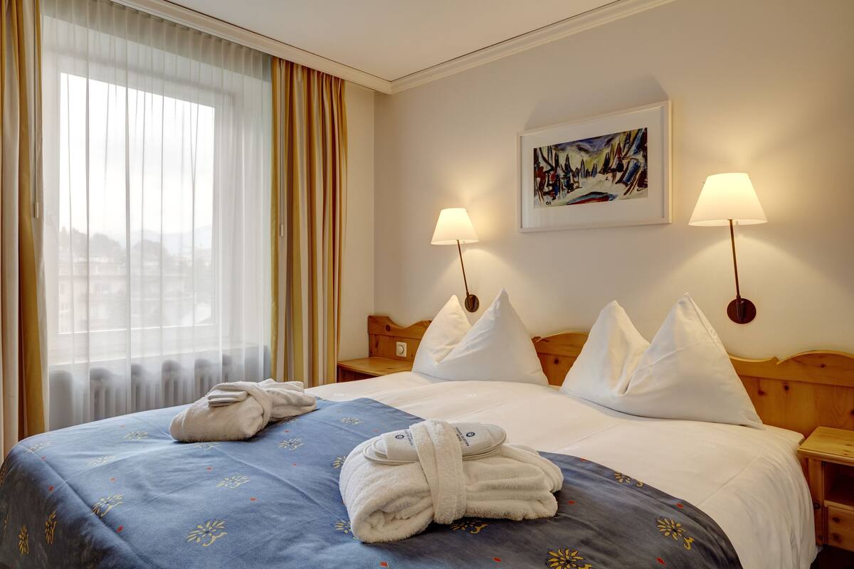 A hotel room with a large bed, two bedside lamps, a painting on the wall, and a window with curtains.