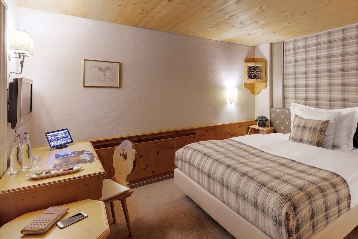 Ett hotellrum med ett träpanelat tak, en säng med ett rutigt överkast, ett skrivbord med en dator och en telefon samt ett litet bord med en växt och en flaska.