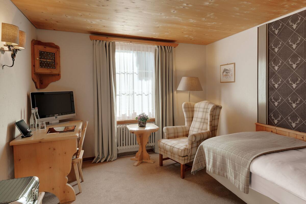 Ett hotellrum med en säng, ett skrivbord med en dator, en stol, en lampa och ett fönster med gardiner.
