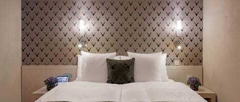 Una cama cuidadosamente tendida con una colcha de cuadros verdes, dos almohadas blancas y una almohada verde con estampado de ciervo. La habitación tiene papel tapiz estampado con motivos de ciervo y dos lámparas fijadas a la pared.