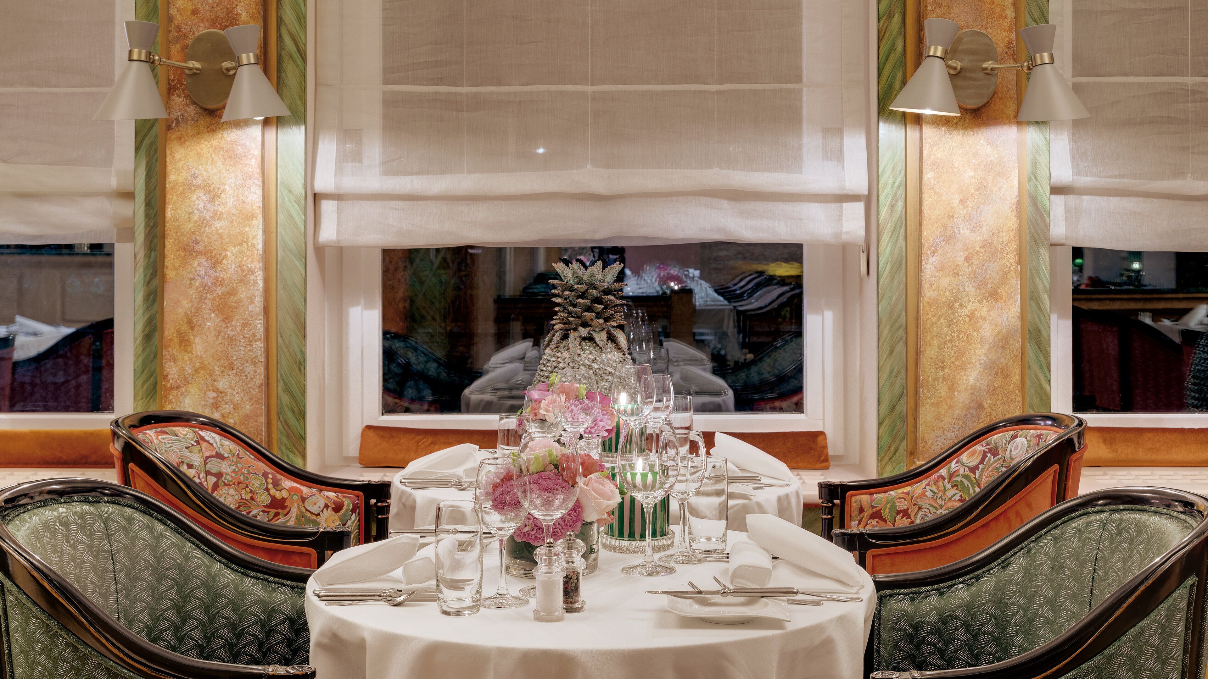 A dining table set with fine china, glassware, and floral centerpieces, surrounded by upholstered chairs with a patterned design.