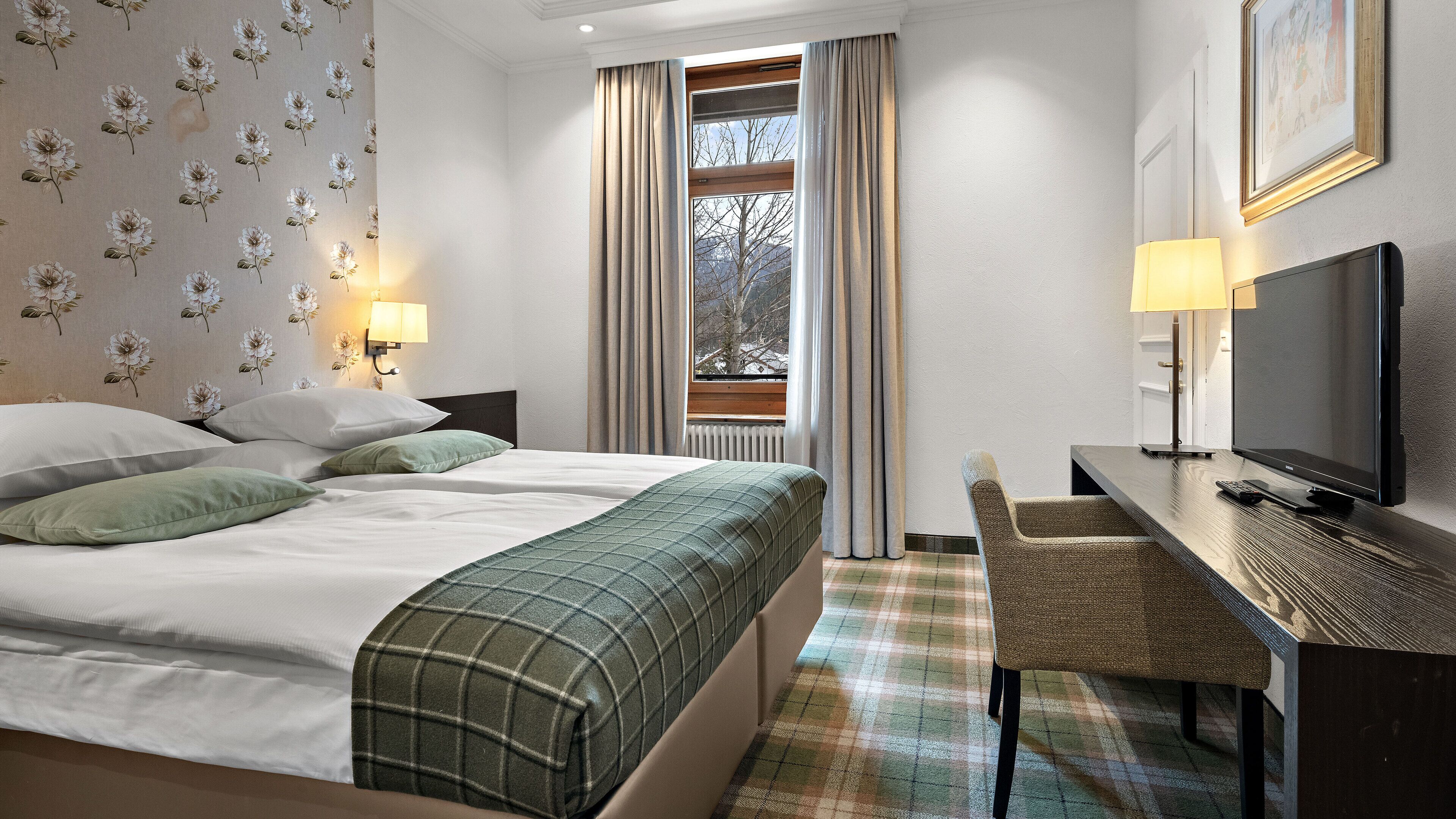 A hotel room with a bed, a desk with a television, a chair, and a window with curtains.