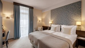 A hotel room with a large bed, two bedside lamps, a headboard with a floral pattern, a desk with a chair, and a view of the outdoors through sheer curtains. - Morosani Schweizerhof (Davos)
