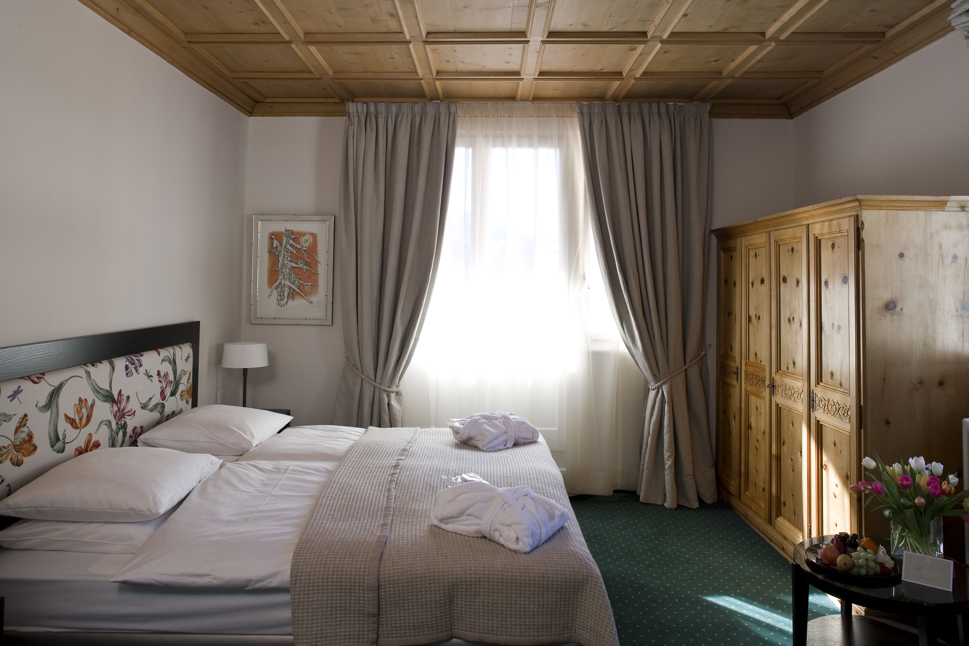 A hotel room with a large bed, a wooden wardrobe, a floral headboard, and a window with curtains.