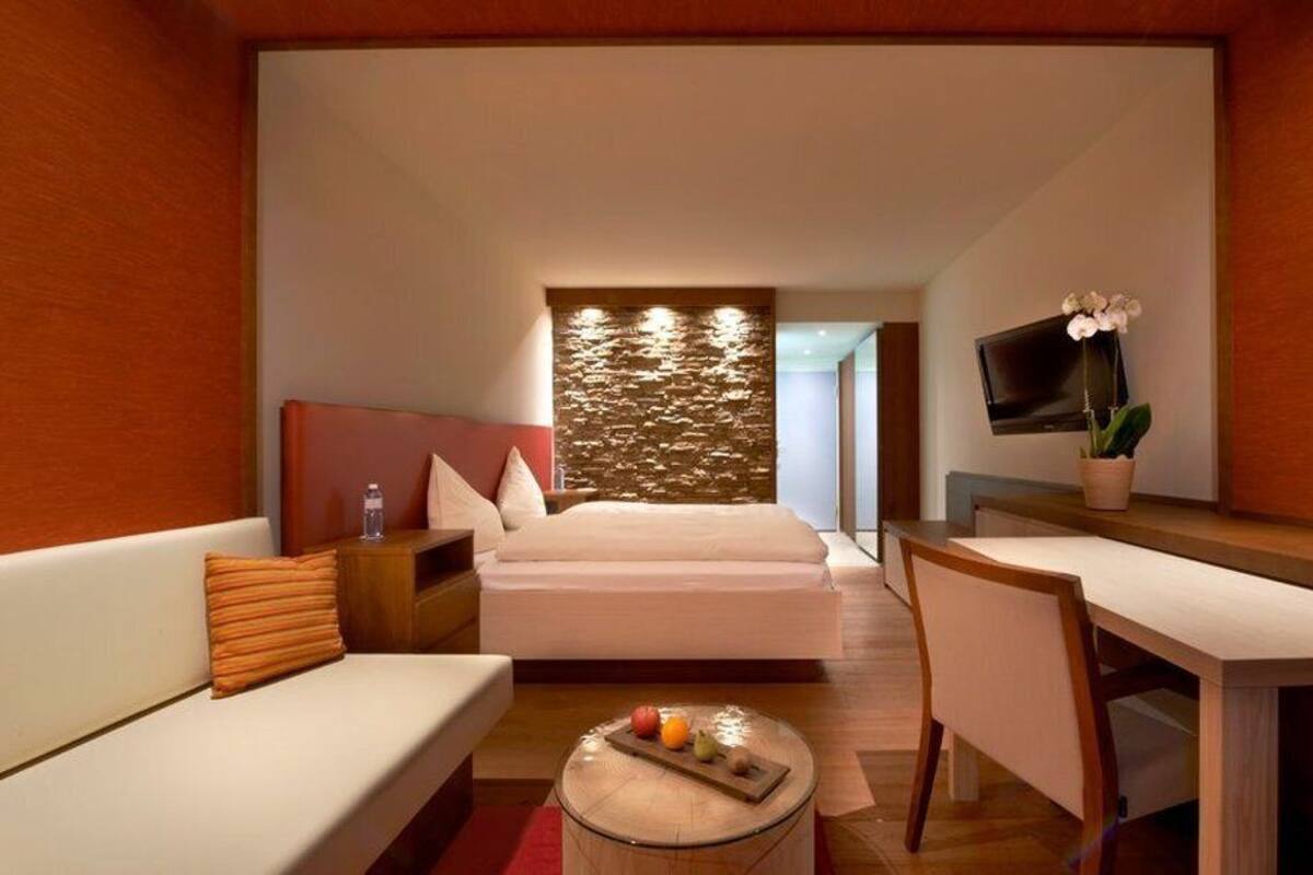 Ein modernes Hotelzimmer mit Bett, Schreibtisch, Stuhl, einem kleinen Tisch mit Früchten, Fernseher und einer Pflanze.