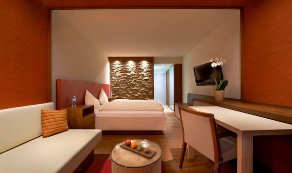 A modern hotel room with a bed, a desk, a chair, a small table with fruit, a TV, and a plant.