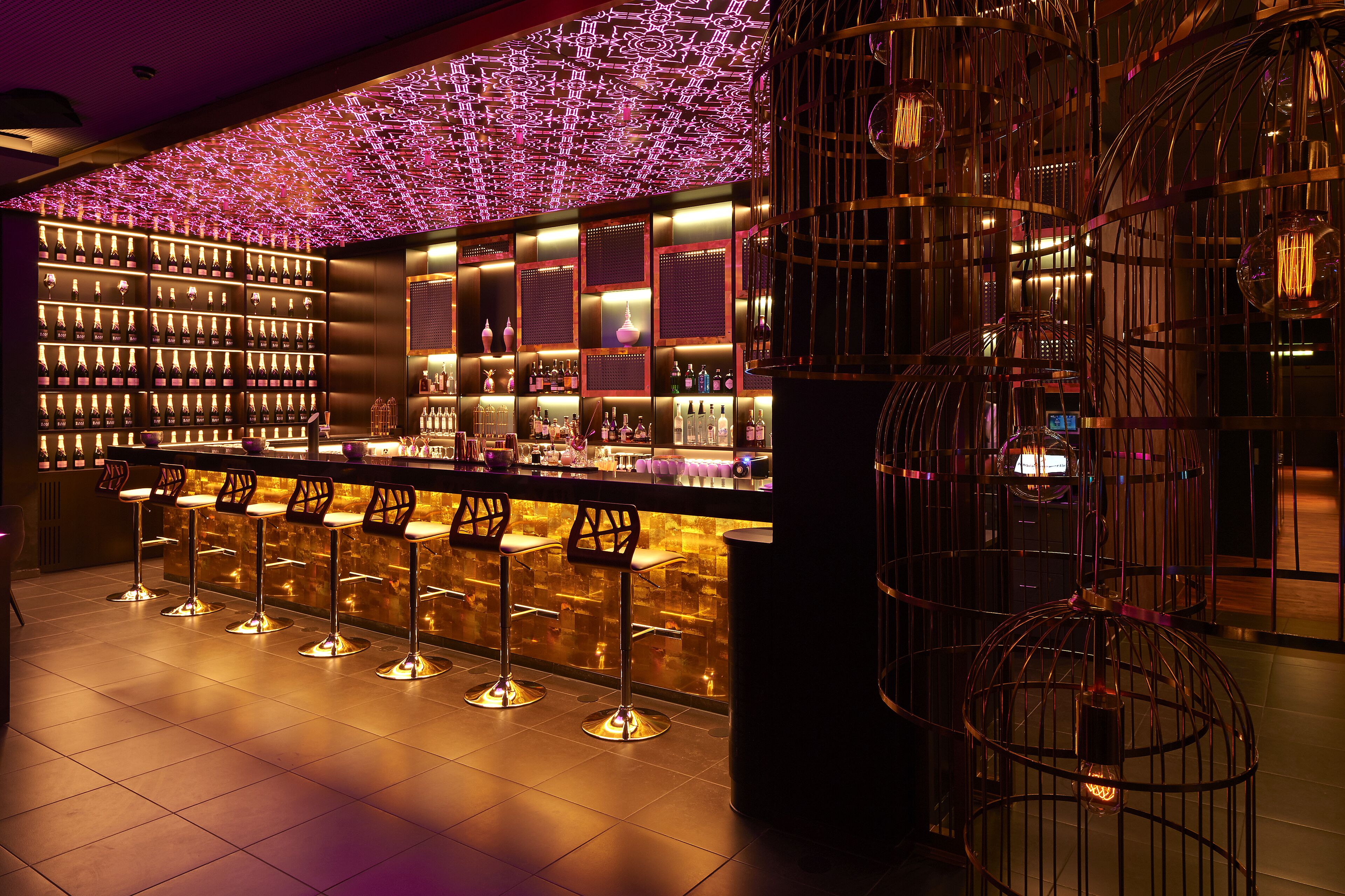 A bar area with multiple stools, a well-lit shelf with bottles, and decorative hanging lights.