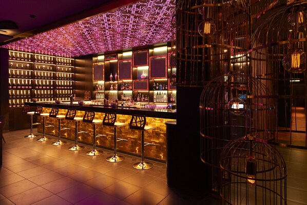A bar area with multiple stools, a well-lit shelf with bottles, and decorative hanging lights.