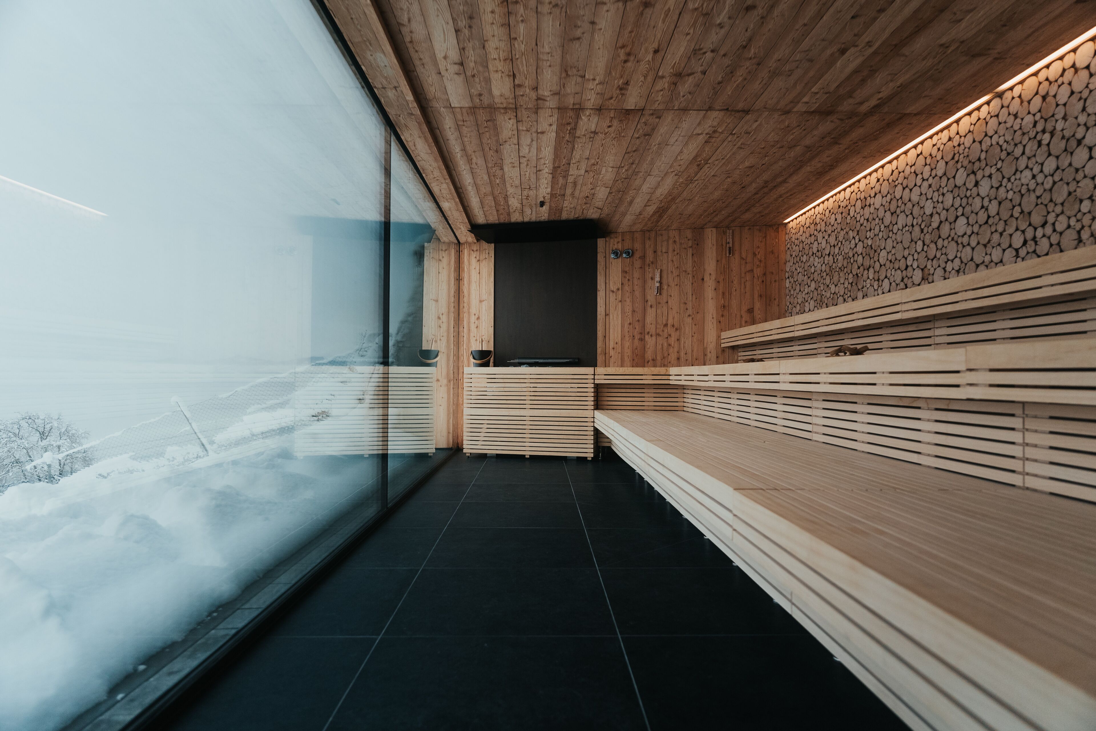 A modern interior with wooden paneling, a black tiled floor, and a view of a snowy landscape through a large glass window.