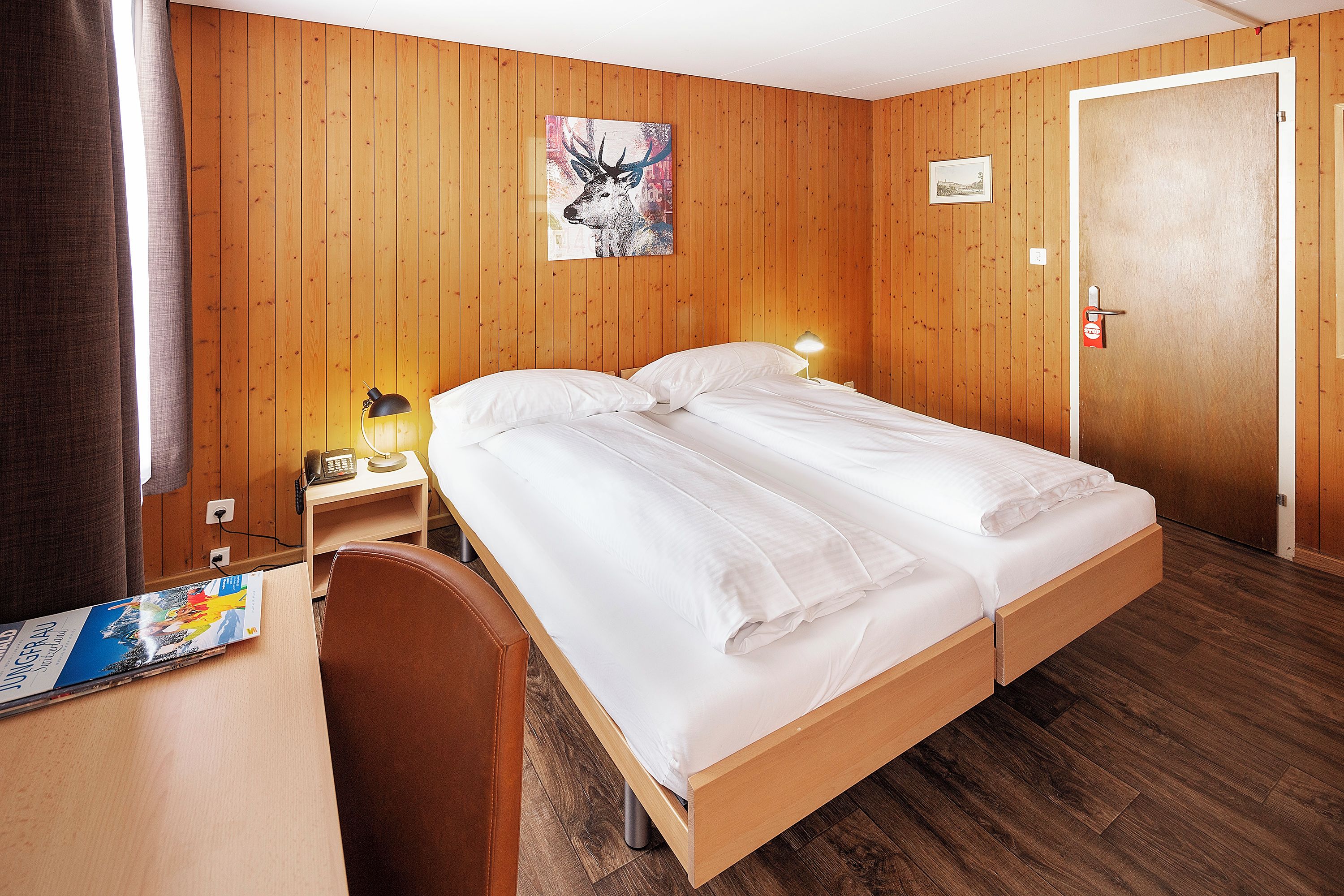 Une chambre d’hôtel avec un mur lambrissé en bois, un lit avec du linge de maison blanc, un bureau avec une lampe et une porte sur laquelle est apposé un panneau indiquant « STOP ».