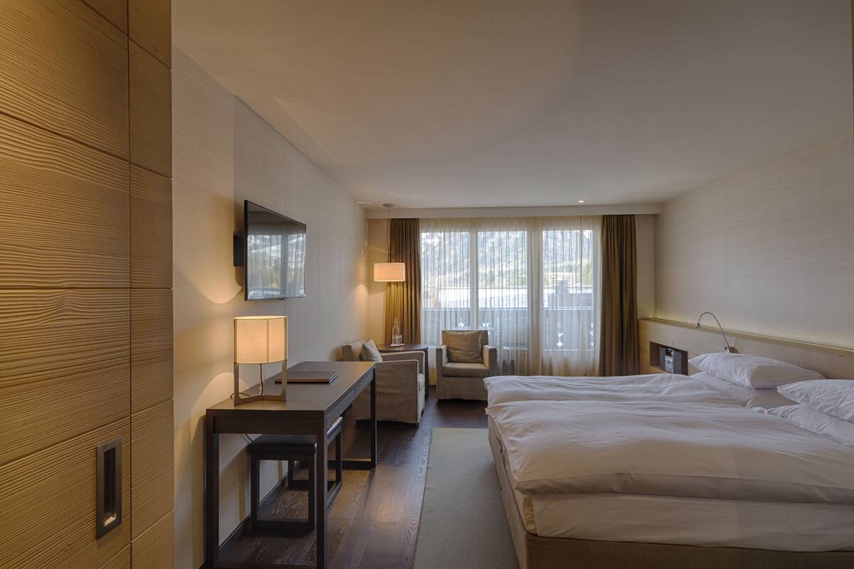 Una habitación de hotel con cama, escritorio, silla, televisor y ventana con cortinas.