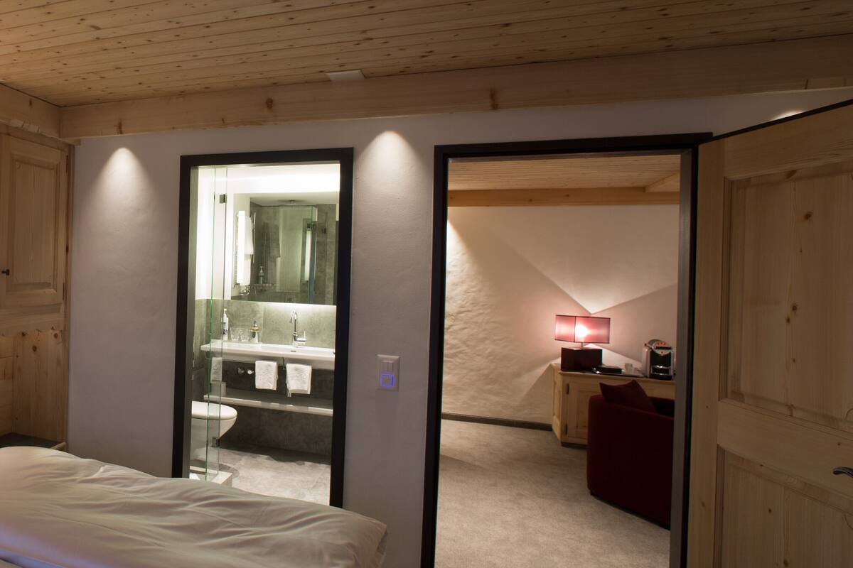 Un dormitorio con cama, un baño con espejo y una pequeña zona de estar con lámpara.