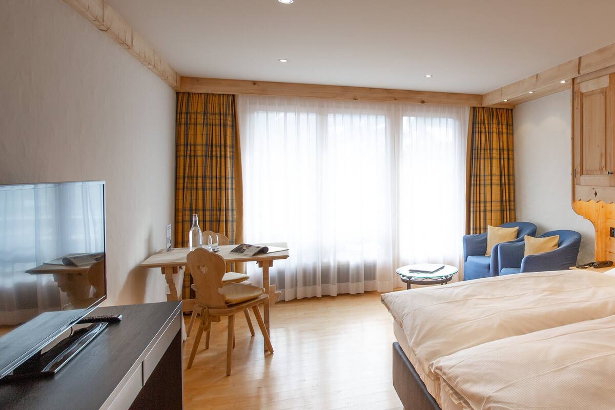 Una habitación de hotel con cama, escritorio con silla, televisor y ventana con cortinas.