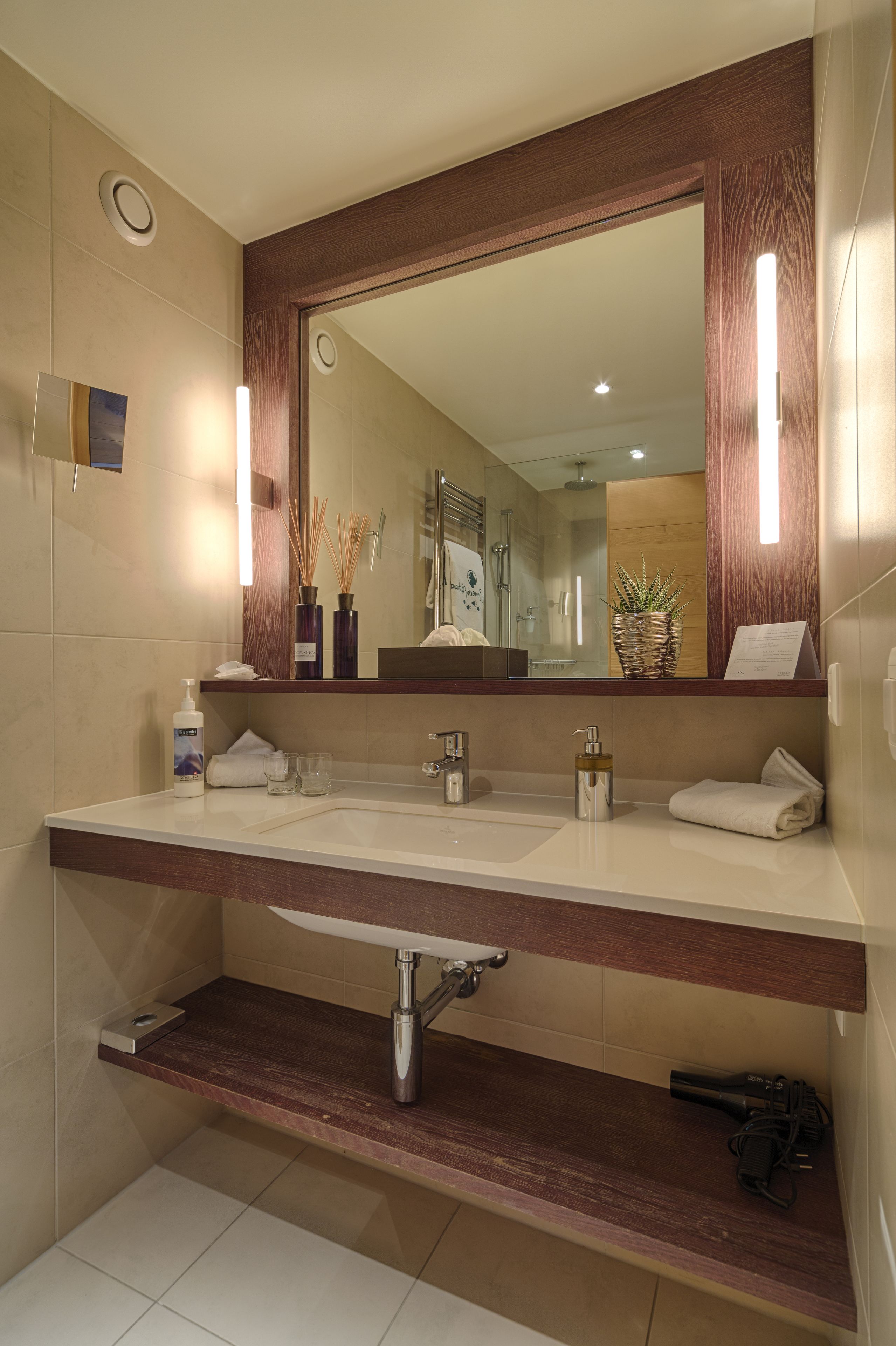 Un baño moderno con un espejo grande, dos lavamanos, un perchero para toallas y un estante con artículos de aseo.
