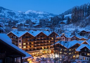 Un complejo turístico en la montaña cubierto de nieve, con varios edificios de madera, incluyendo un hotel con balcones y una entrada iluminada.