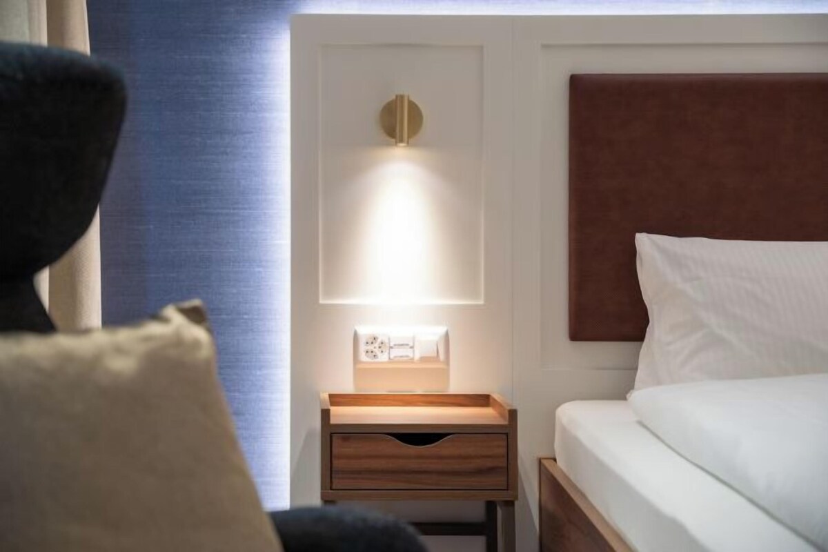 A hotel room with a bed, a bedside table with a lamp, and a small drawer unit.