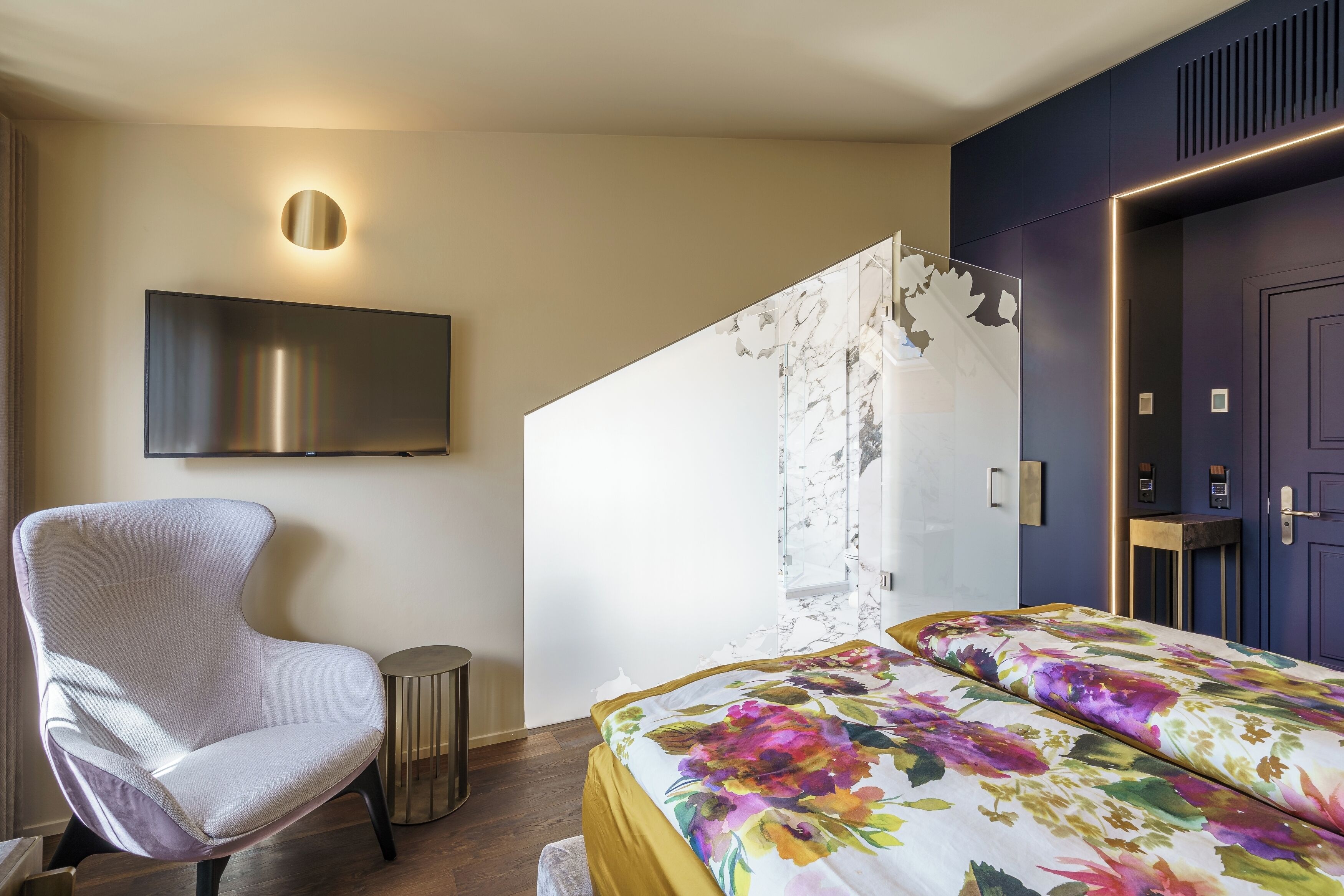 A modern hotel room with a large bed, a comfortable armchair, a flat-screen TV mounted on the wall, and a unique marble-patterned wall.