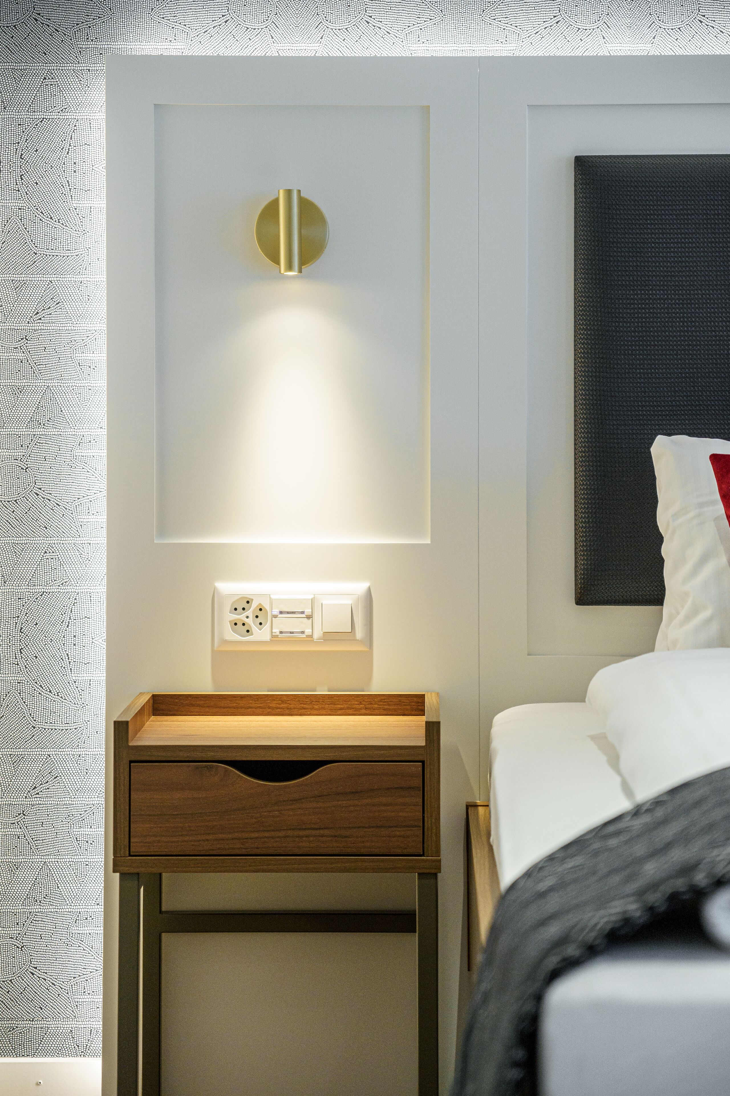 A hotel room with a bedside table, a wall-mounted light, a power outlet, and a bed with a pillow.
