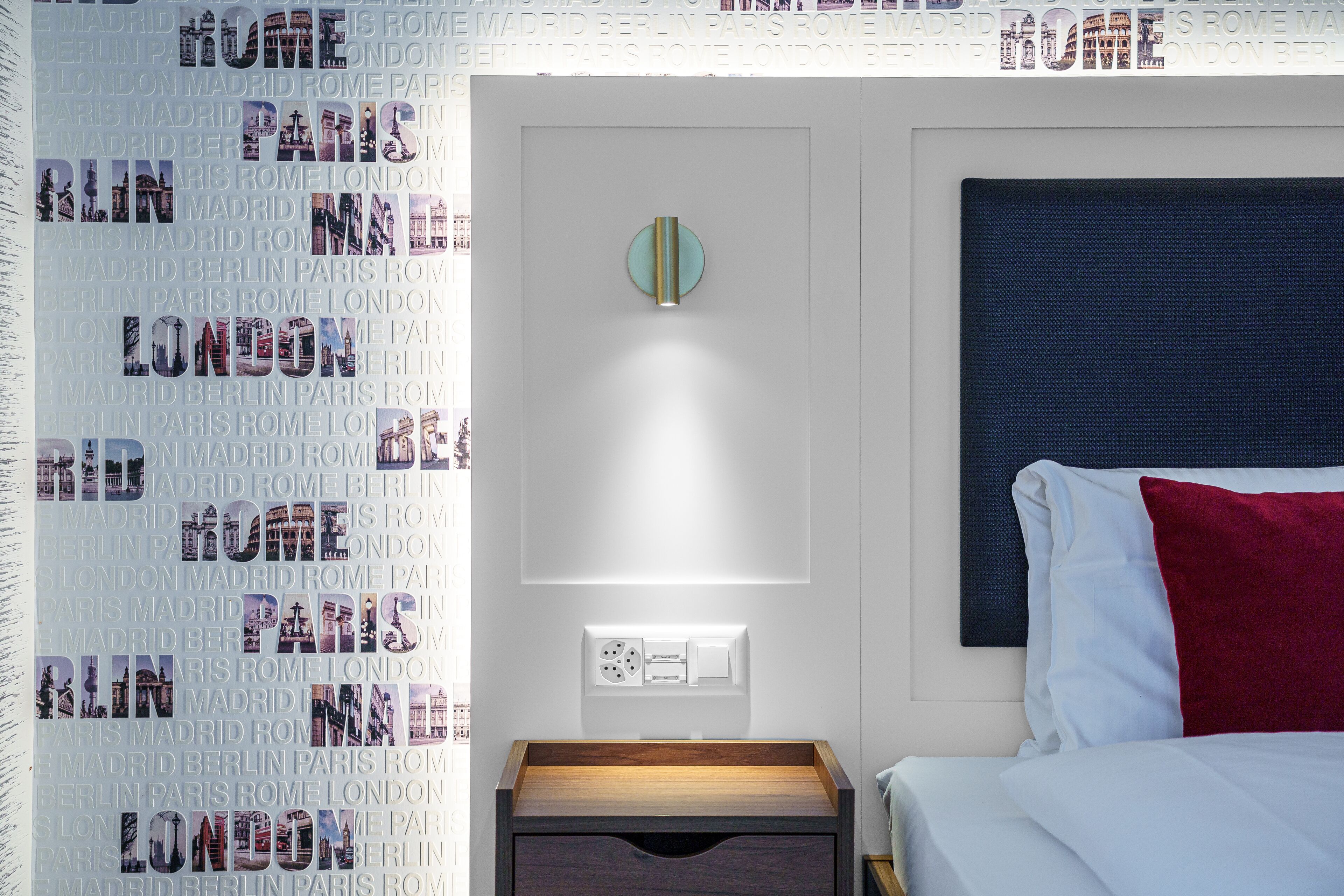 A hotel room with a bed, a nightstand, and a wall with repeated city names.