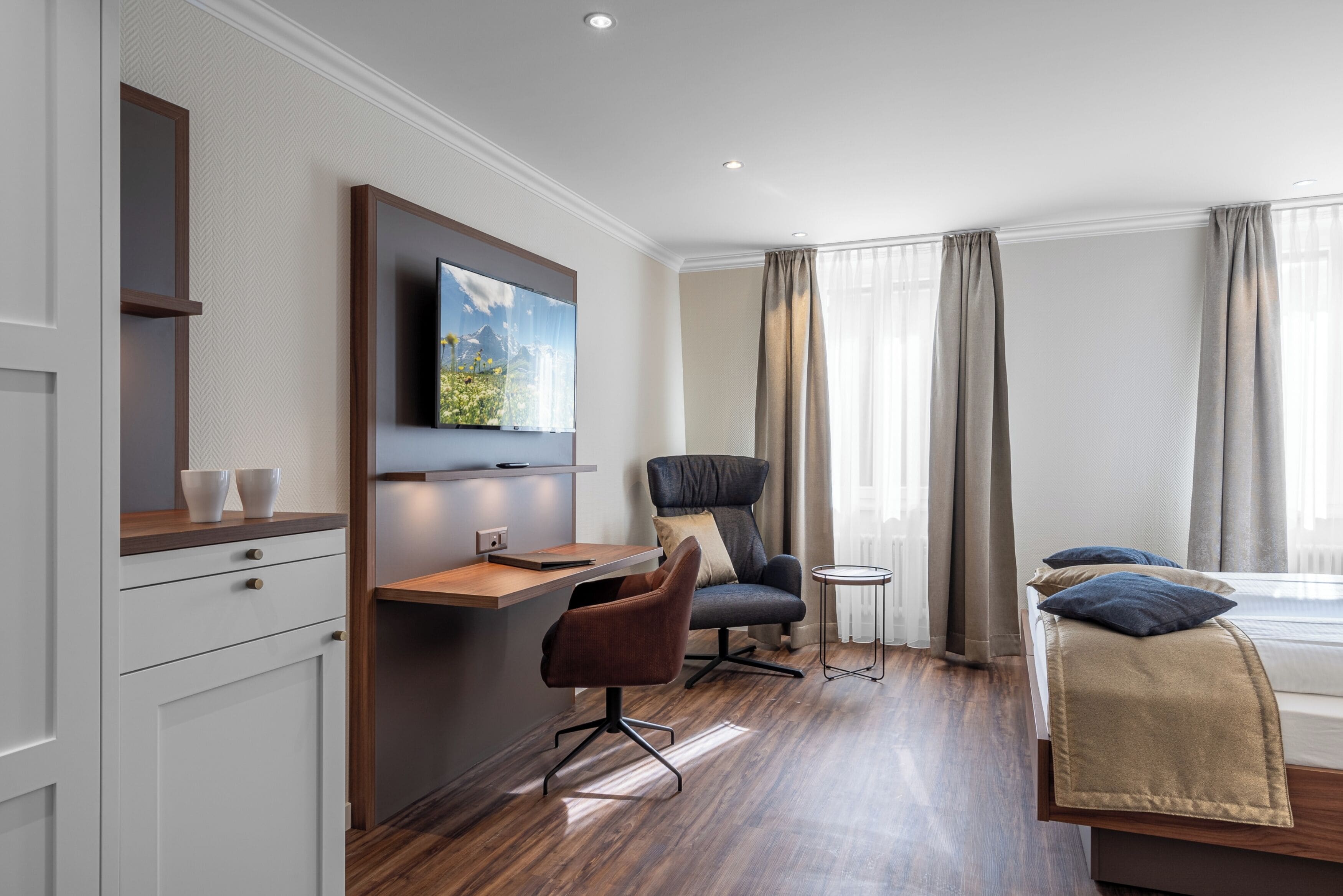 A modern hotel room with a wooden desk, a comfortable armchair, a bed with a beige blanket, and a large window with curtains.