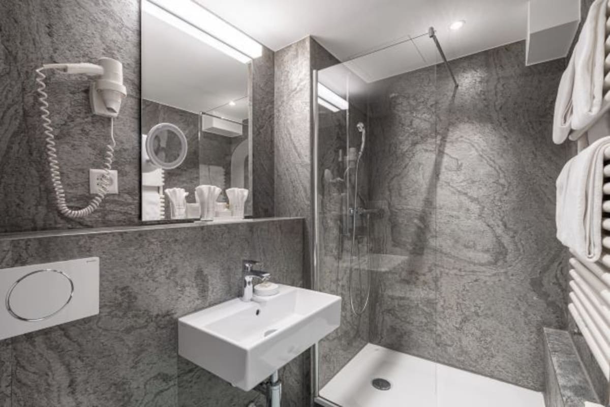 A modern bathroom with a marble wall, a glass shower enclosure, a white sink, and a hairdryer mounted on the wall.