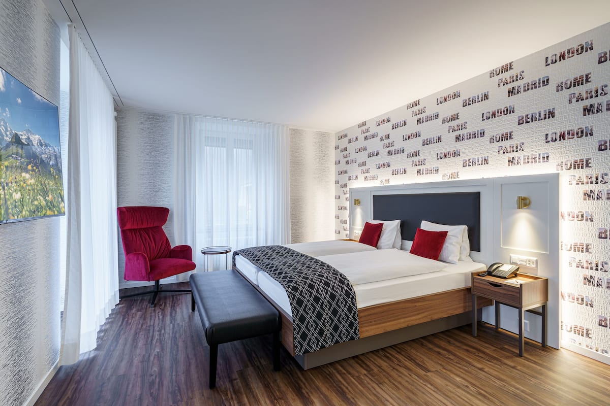 A modern hotel room with a large bed, a red chair, a small table, and a wall with city names.