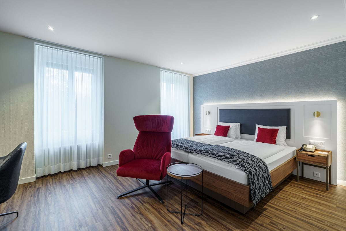 A modern hotel room with a large bed, a red armchair, a small side table, and a wooden floor.