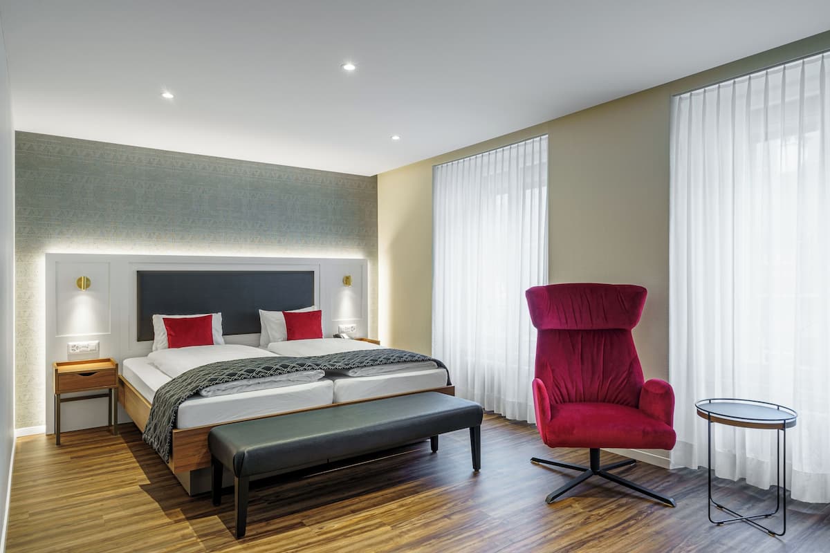 A modern hotel room with a large bed, a red armchair, a small table, and a bench.