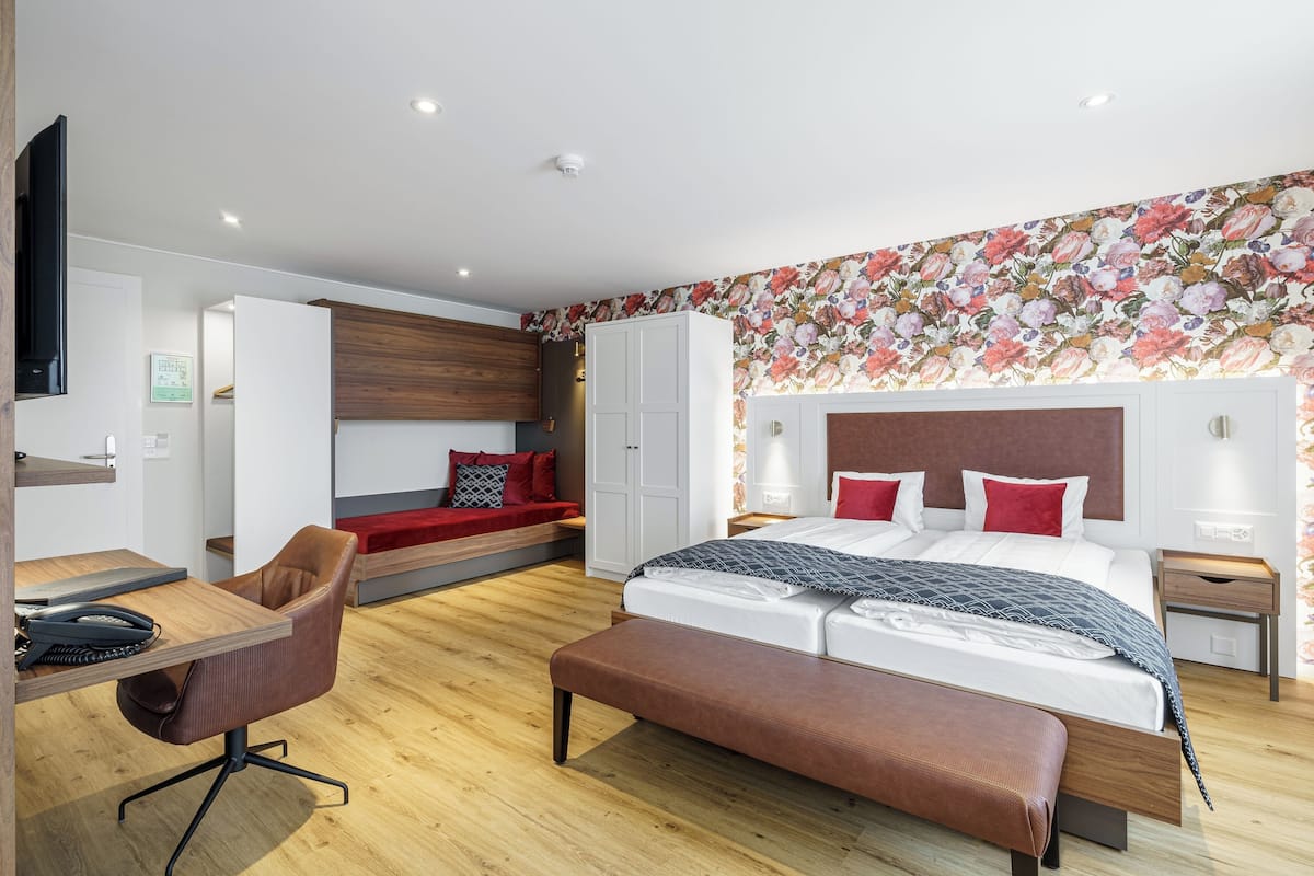 A modern hotel room with a wooden floor, a large bed with red pillows, a desk with a chair, a wardrobe, and a TV mounted on the wall.