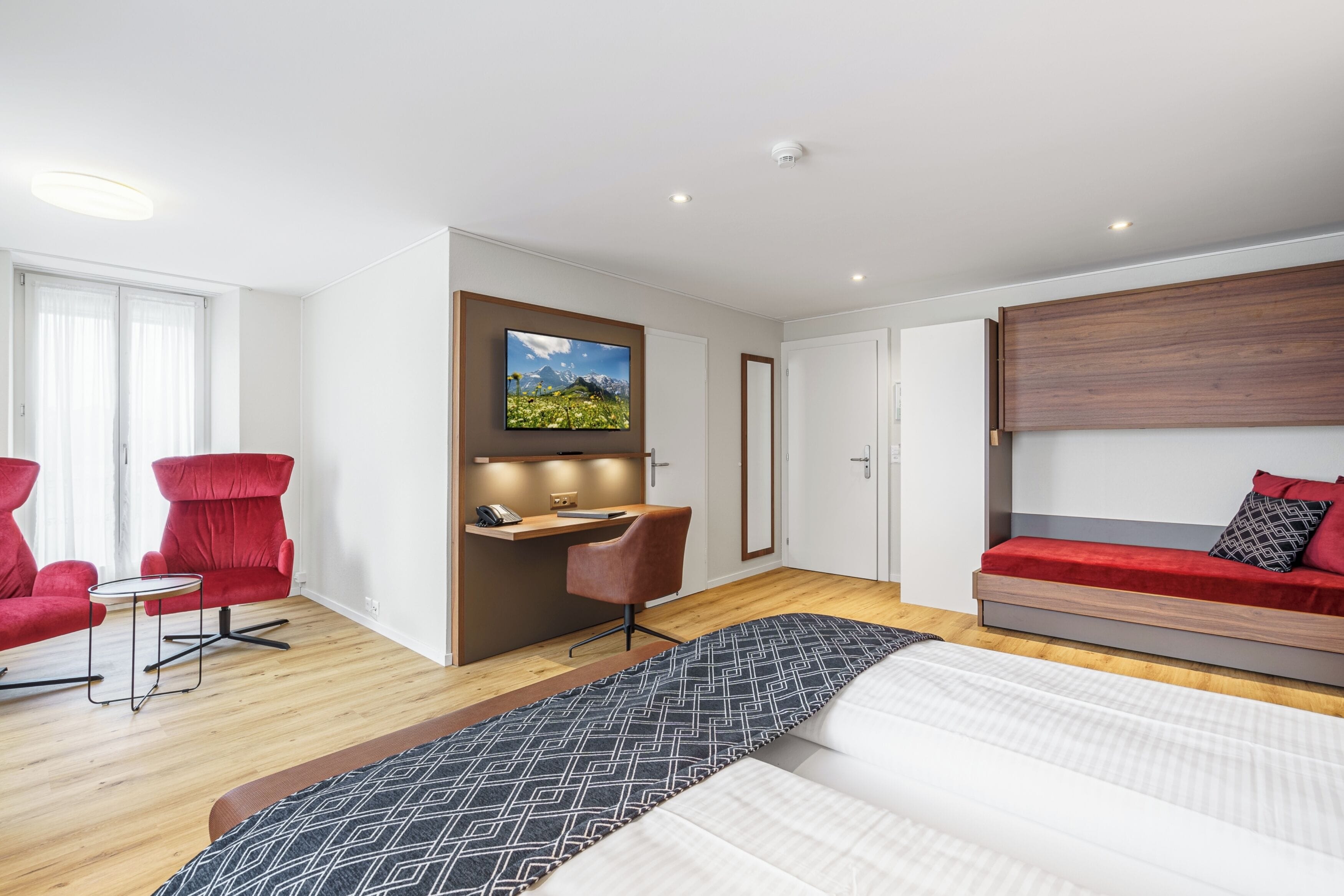 A modern hotel room with a bed, a desk, two red chairs, a TV, and a red sofa.