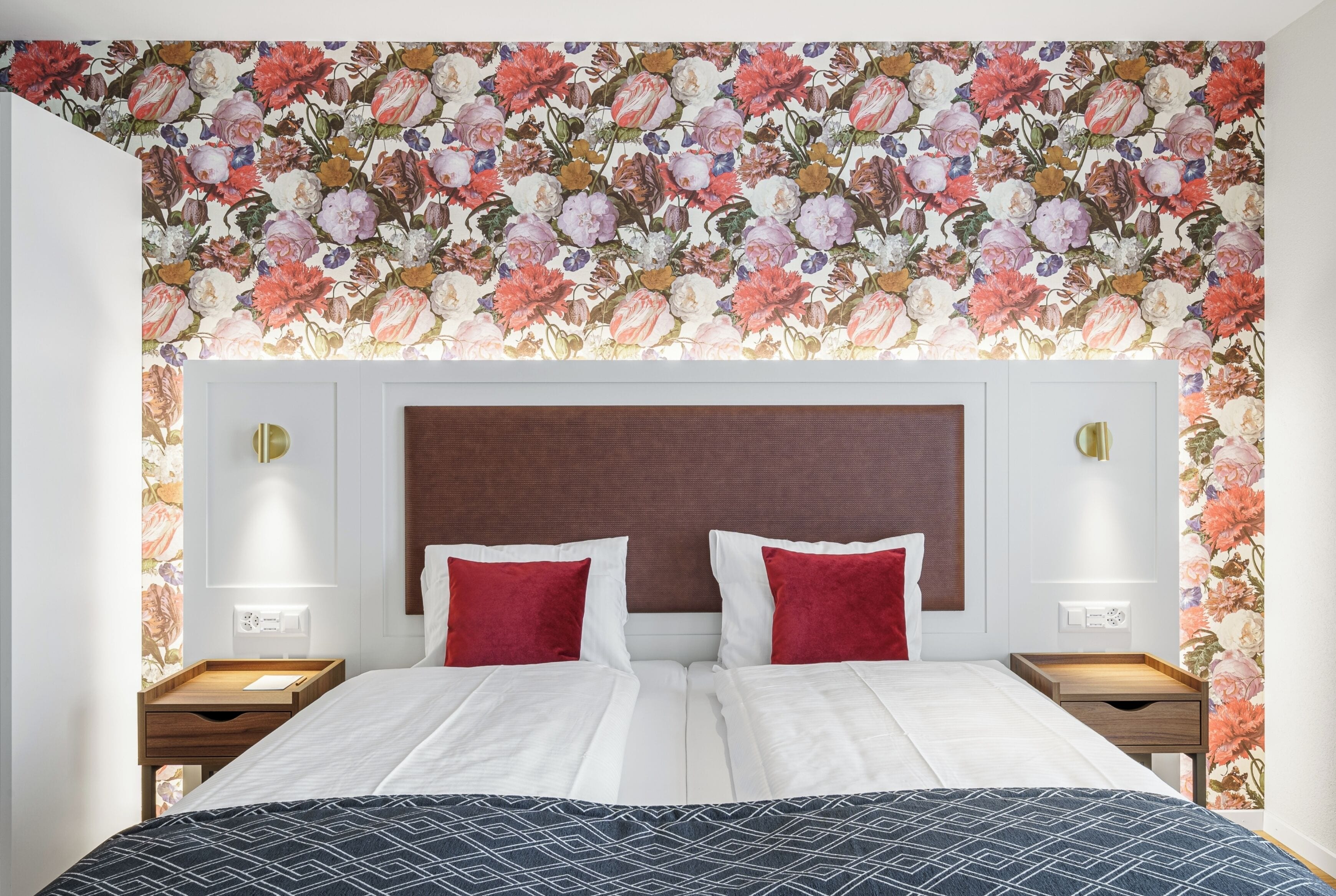 Una cama doble con cabecera estampada, dos mesitas de noche con lámparas y un diseño de papel tapiz floral.