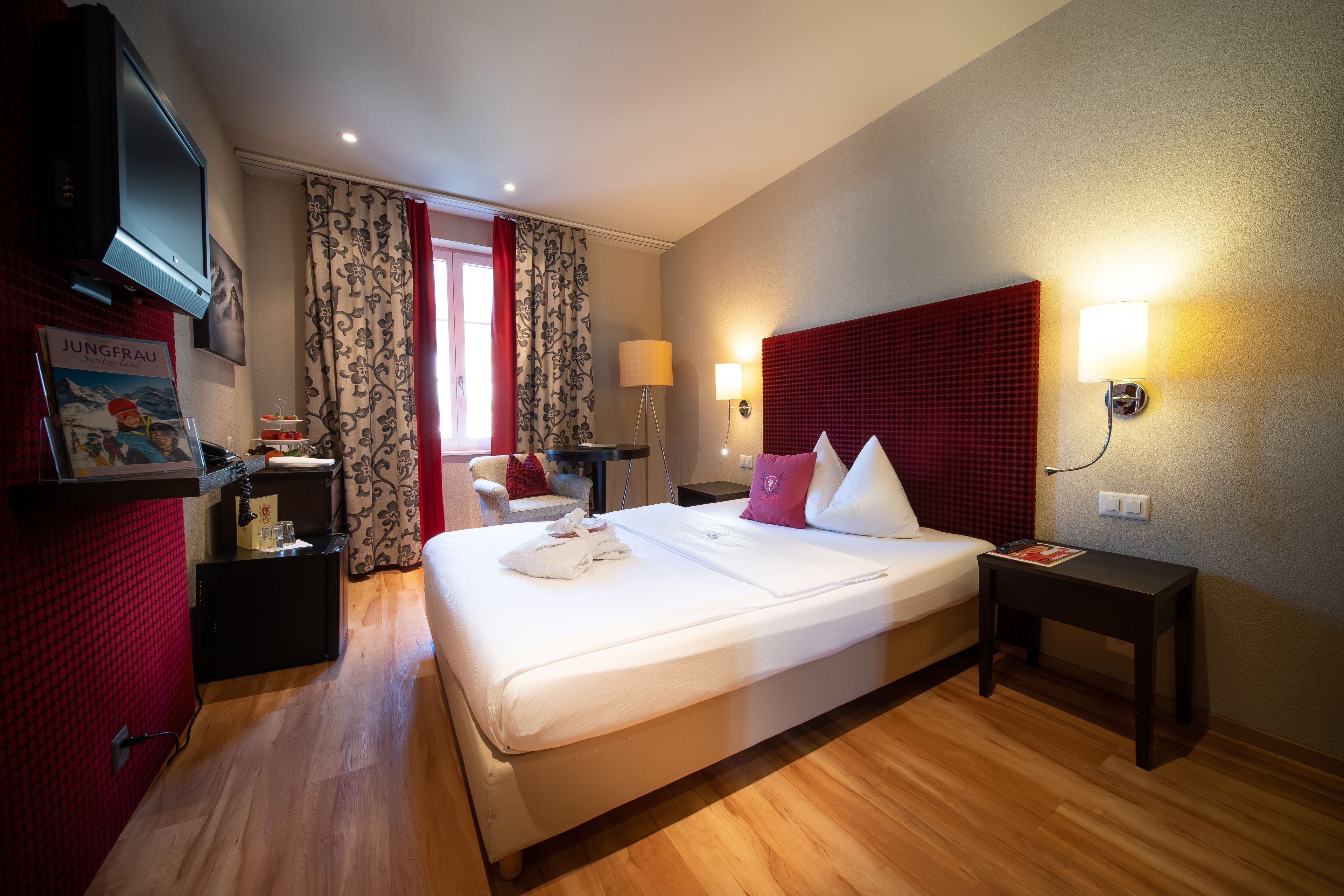 Una habitación de hotel con una cama grande, un televisor montado en la pared, un escritorio con una lámpara y una pared de acento roja.
