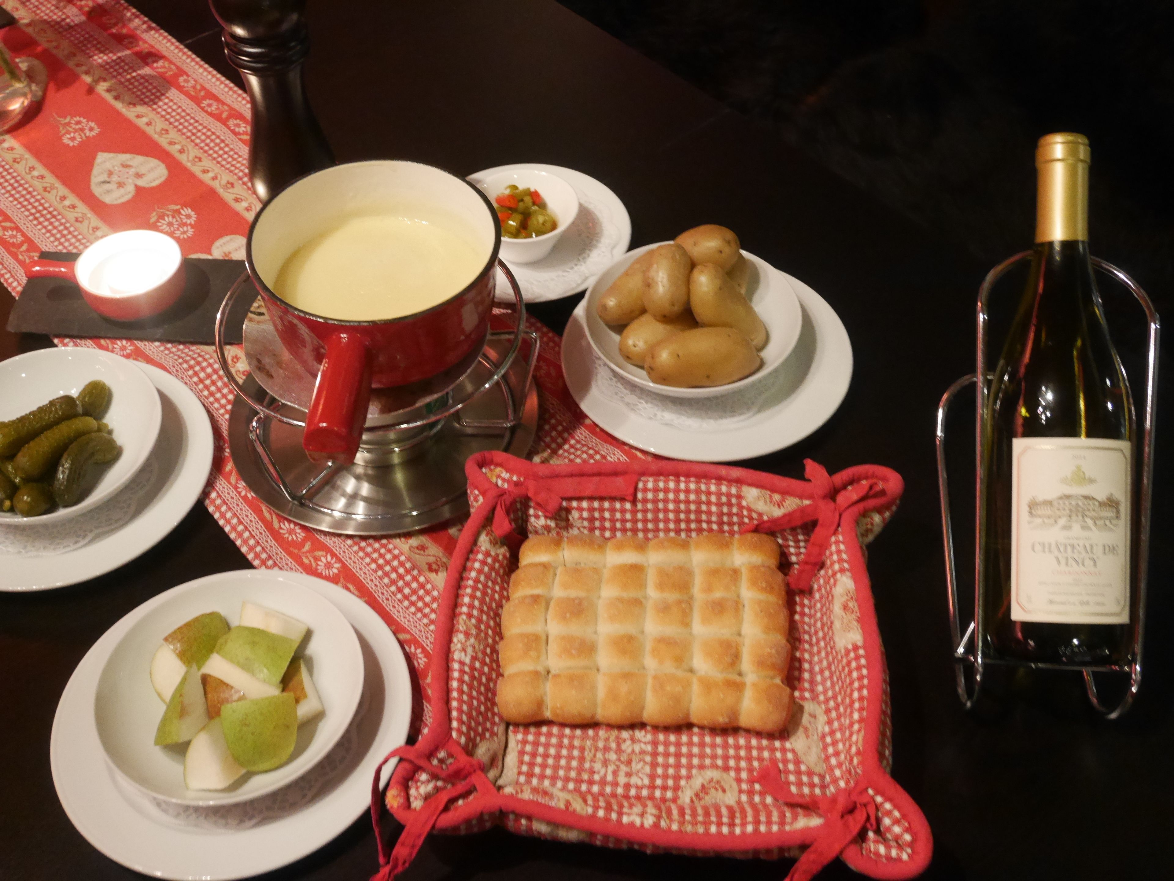 A dining table with various food items, a cheese fondue pot, a bottle of wine, and a basket of bread.